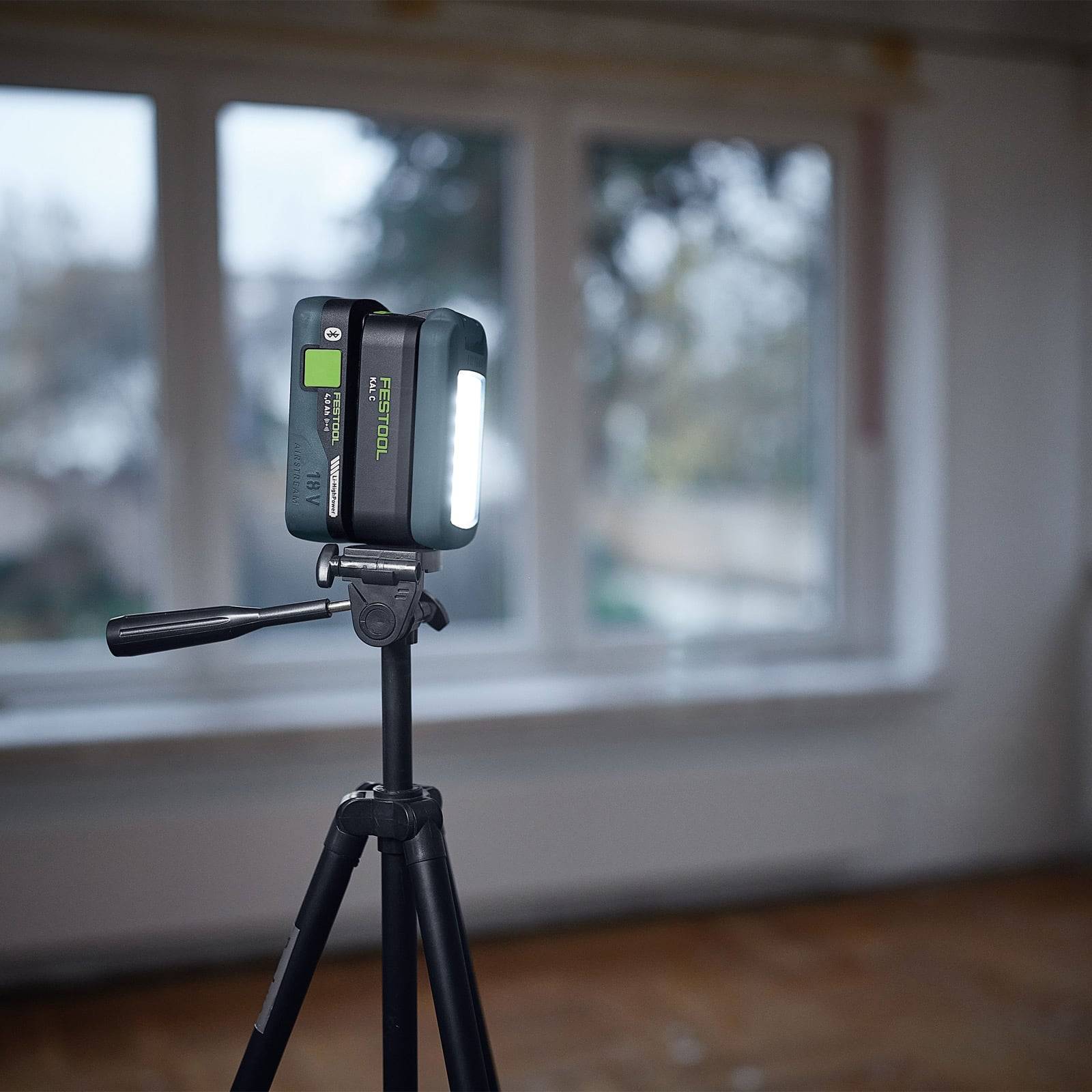 Ein Stativ mit einem montierten Lasernivelliergerät steht vor einer unscharfen Fensteransicht. Das Lasernivelliergerät sendet ein helles Licht aus.