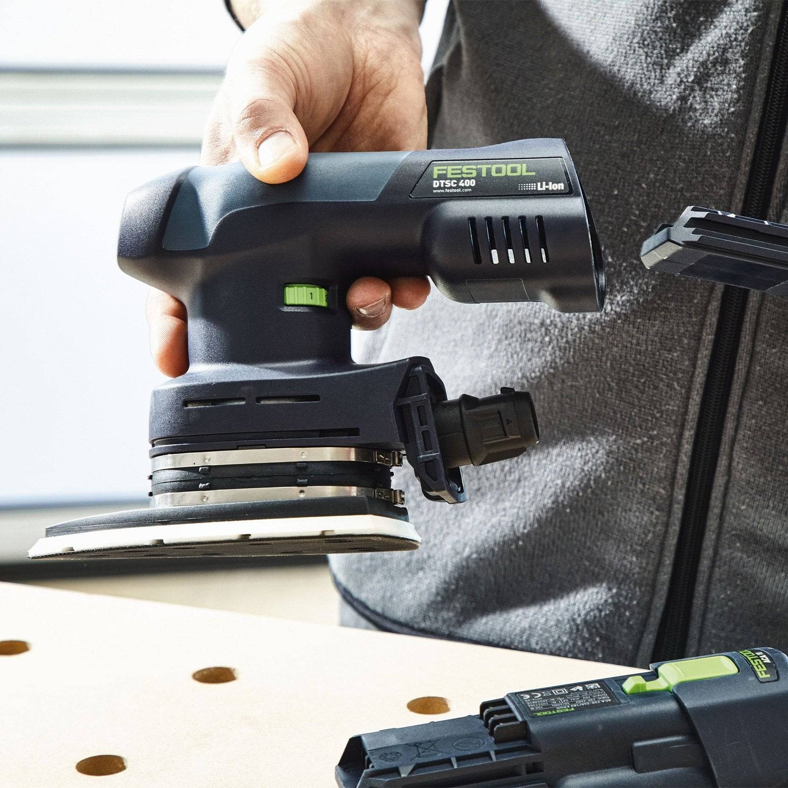 Eine Hand, die einen Festool Exzenterschleifer in der Nähe einer Holzwerkbank hält und das kompakte Design und die präzisen Steuerungsmerkmale des Werkzeugs präsentiert.