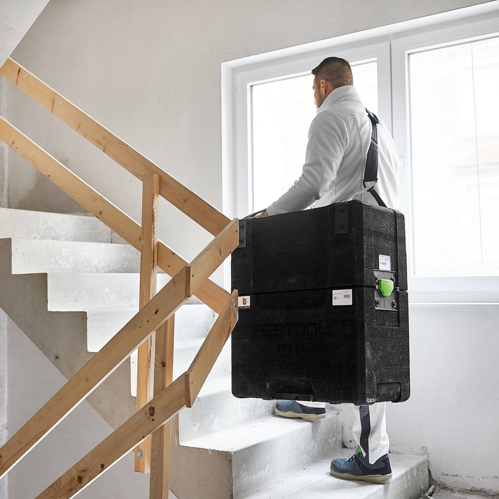 Eine Person trägt einen großen schwarzen Behälter eine Treppe hinauf und trägt dabei eine weiße Jacke und graue Hosen. Sonnenlicht strömt durch ein nahegelegenes Fenster.
