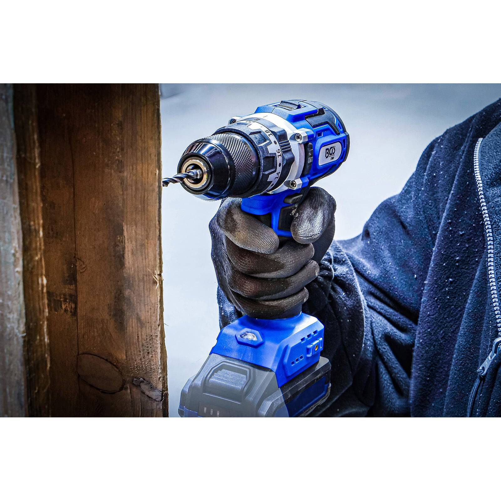 Eine Person trägt eine dunkle Jacke und verwendet eine blaue kabellose Elektrobohrmaschine, um ein Loch in eine Holzoberfläche zu bohren.