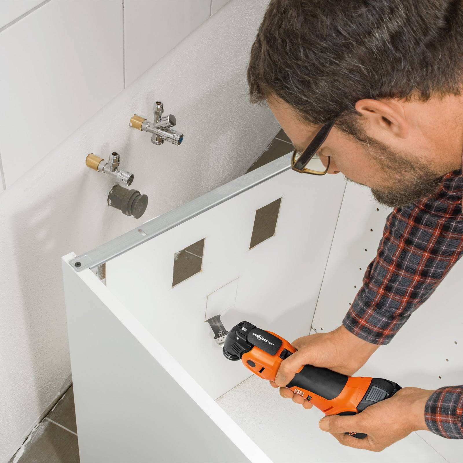 Ein Mann installiert einen weißen Schrank mit einer Bohrmaschine. Der Schrank verfügt über Öffnungen für Sanitärleitungen, und die Wand ist teilweise gefliest.