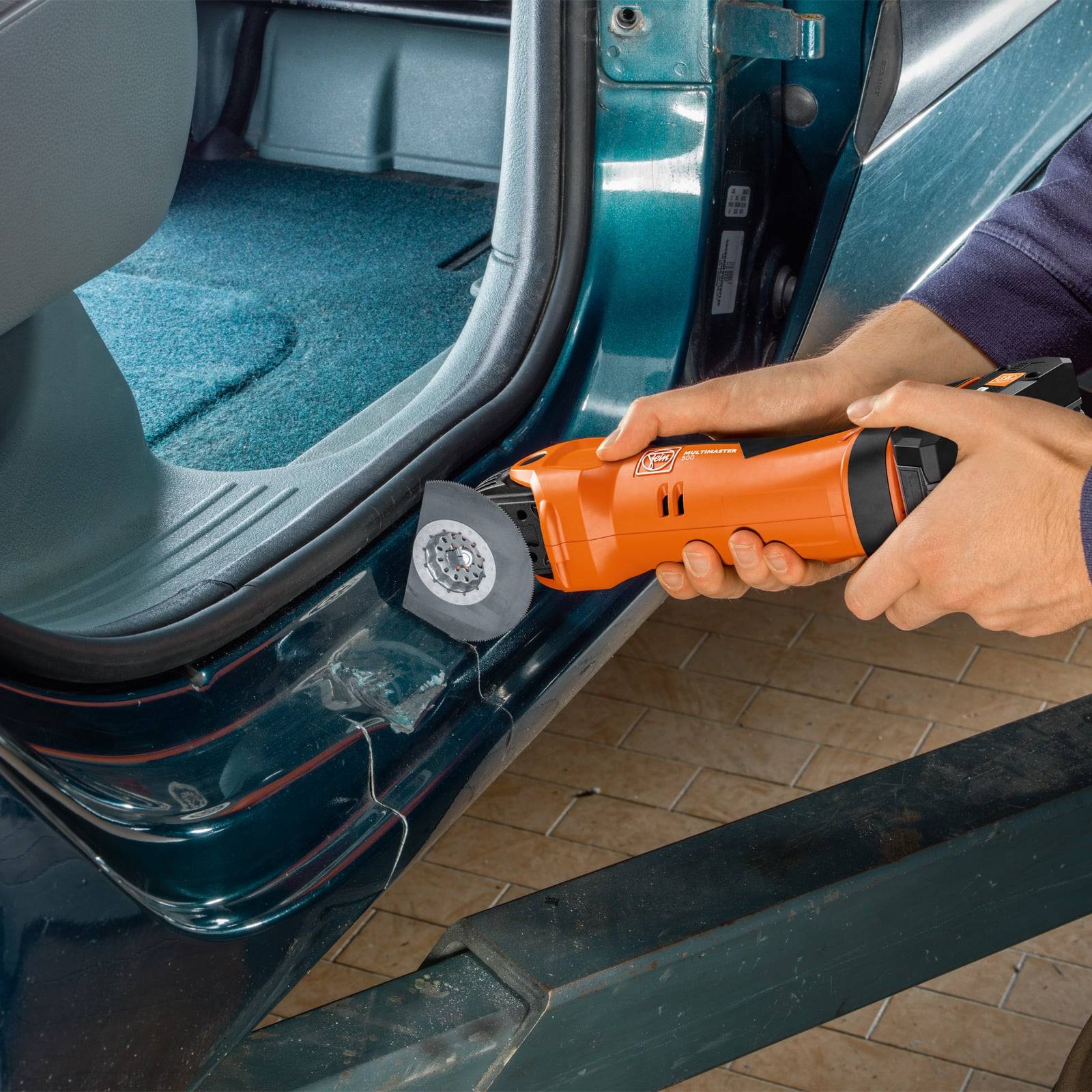 Eine Person verwendet ein oszillierendes Multiwerkzeug, um den unteren Türrahmen eines Fahrzeugs zu schleifen. Das Werkzeug ist orange und hat eine kreisförmige Schleifscheibe.