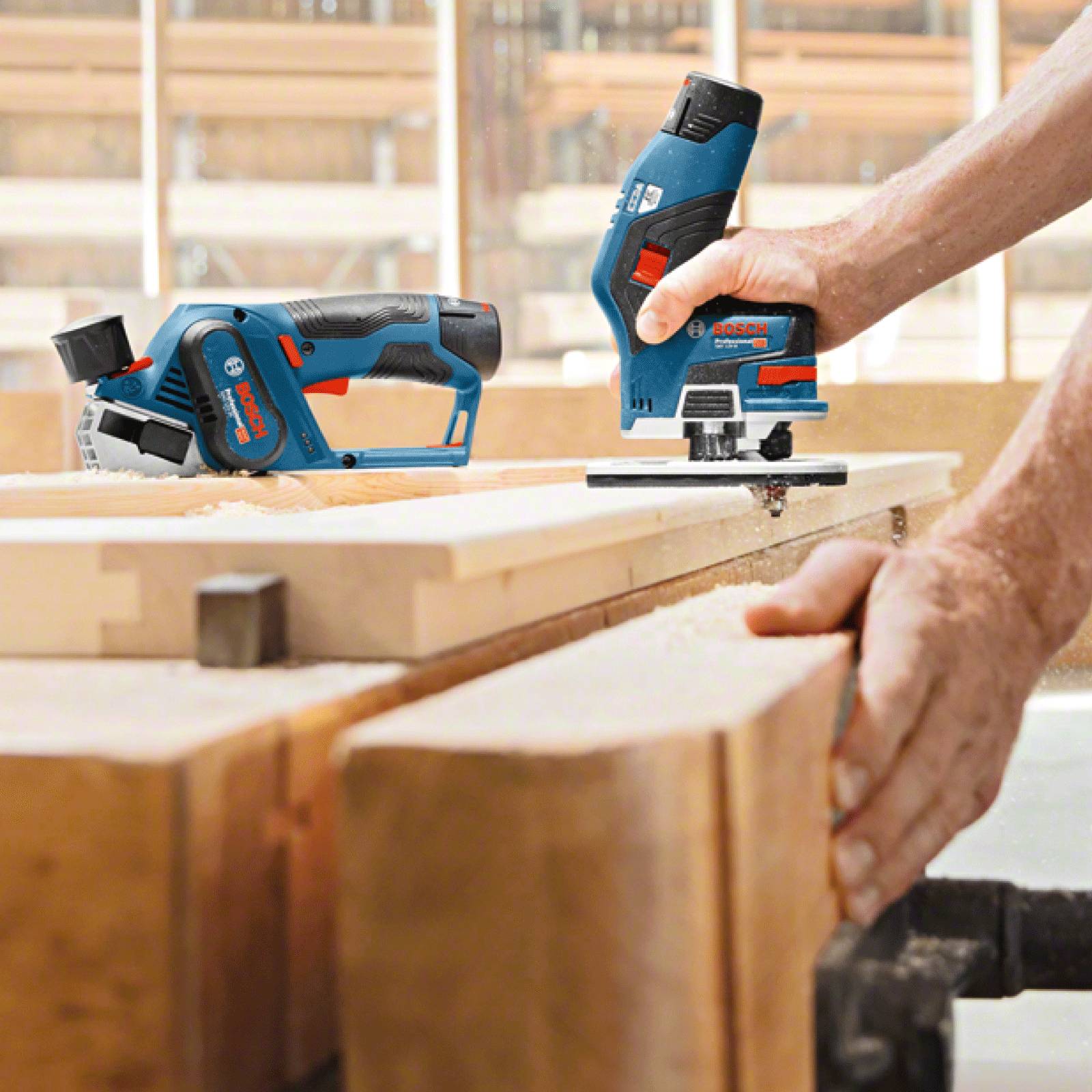 Eine Person bedient einen blau-schwarzen kabellosen elektrischen Hobel auf einem Holzbrett, wobei ein weiteres Werkzeug im Hintergrund auf einer Werkbank zu sehen ist.