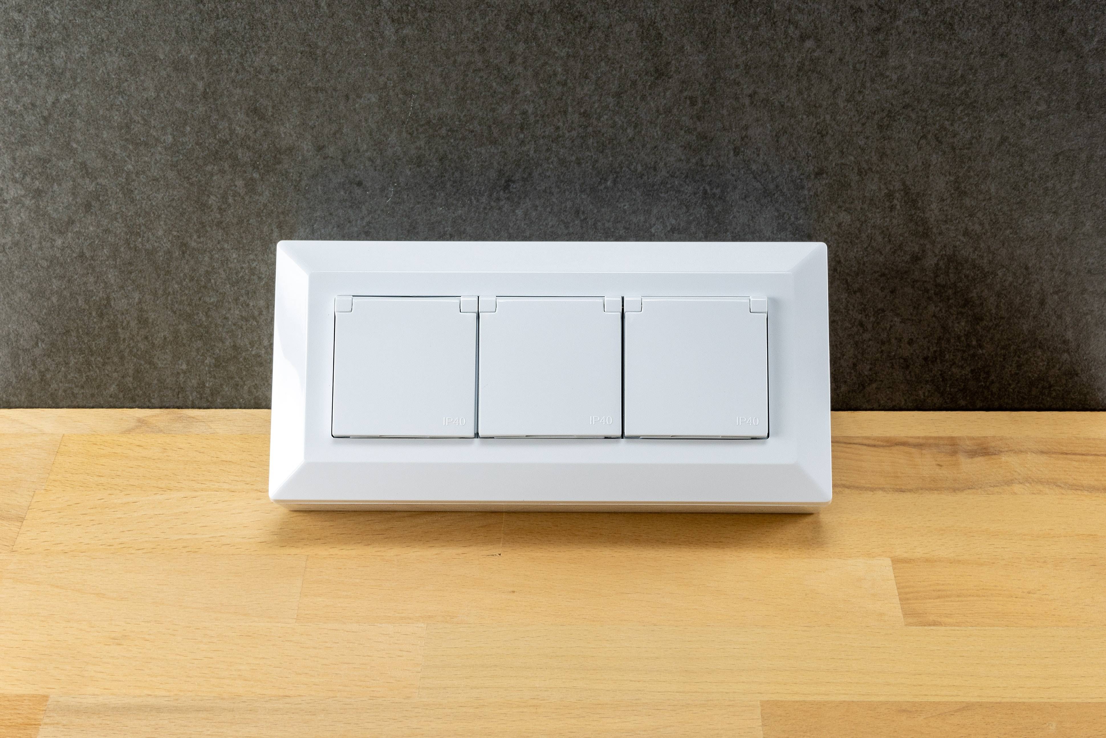 Eine weiße elektrische Verteilerdose mit drei Schaltern, die auf einer Holzoberfläche vor einer grauen Wand montiert ist.