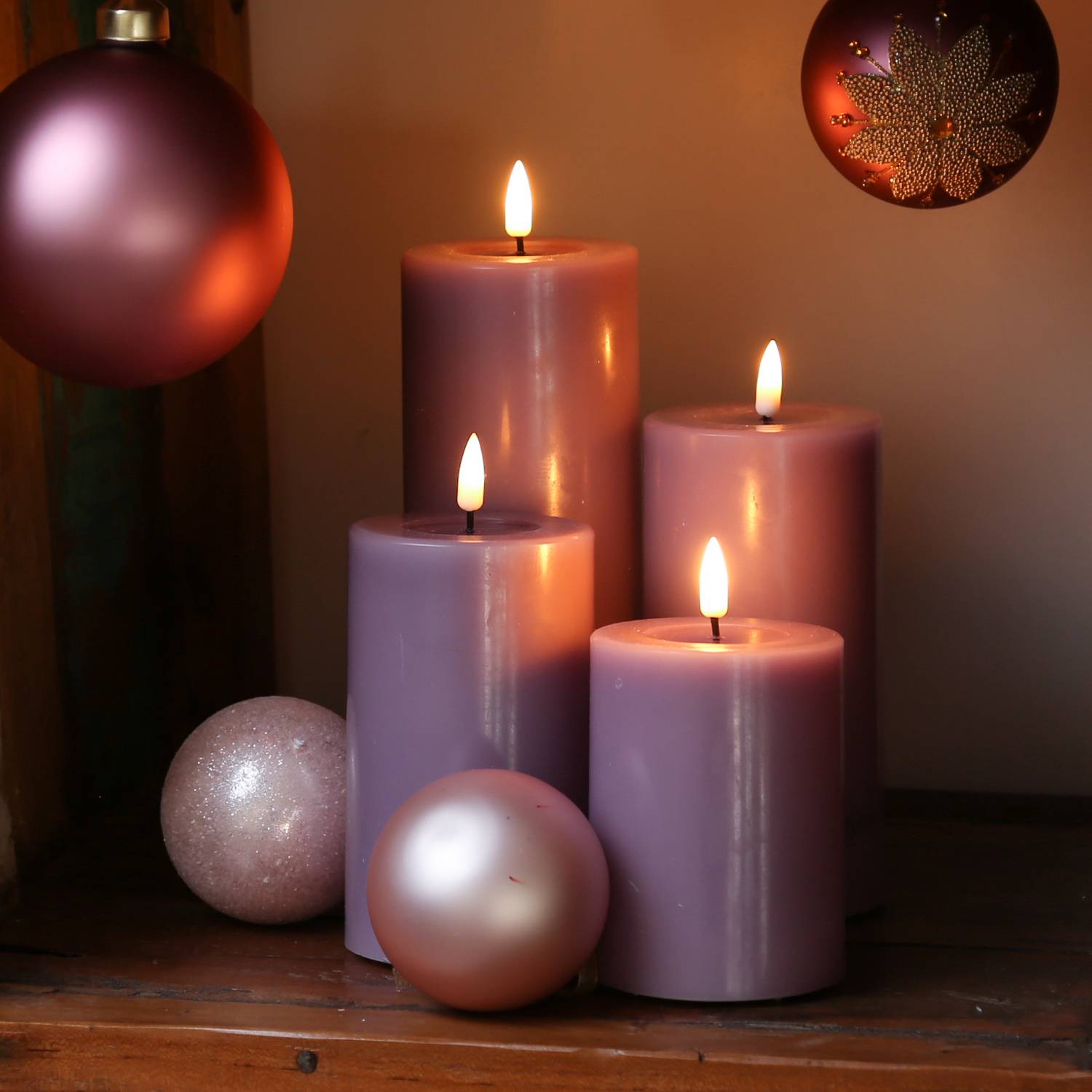 Vier leuchtende lila Kerzen verschiedener Höhen auf einer Holzoberfläche, umgeben von rosa und lila Kugeln, die eine warme, festliche Atmosphäre schaffen.