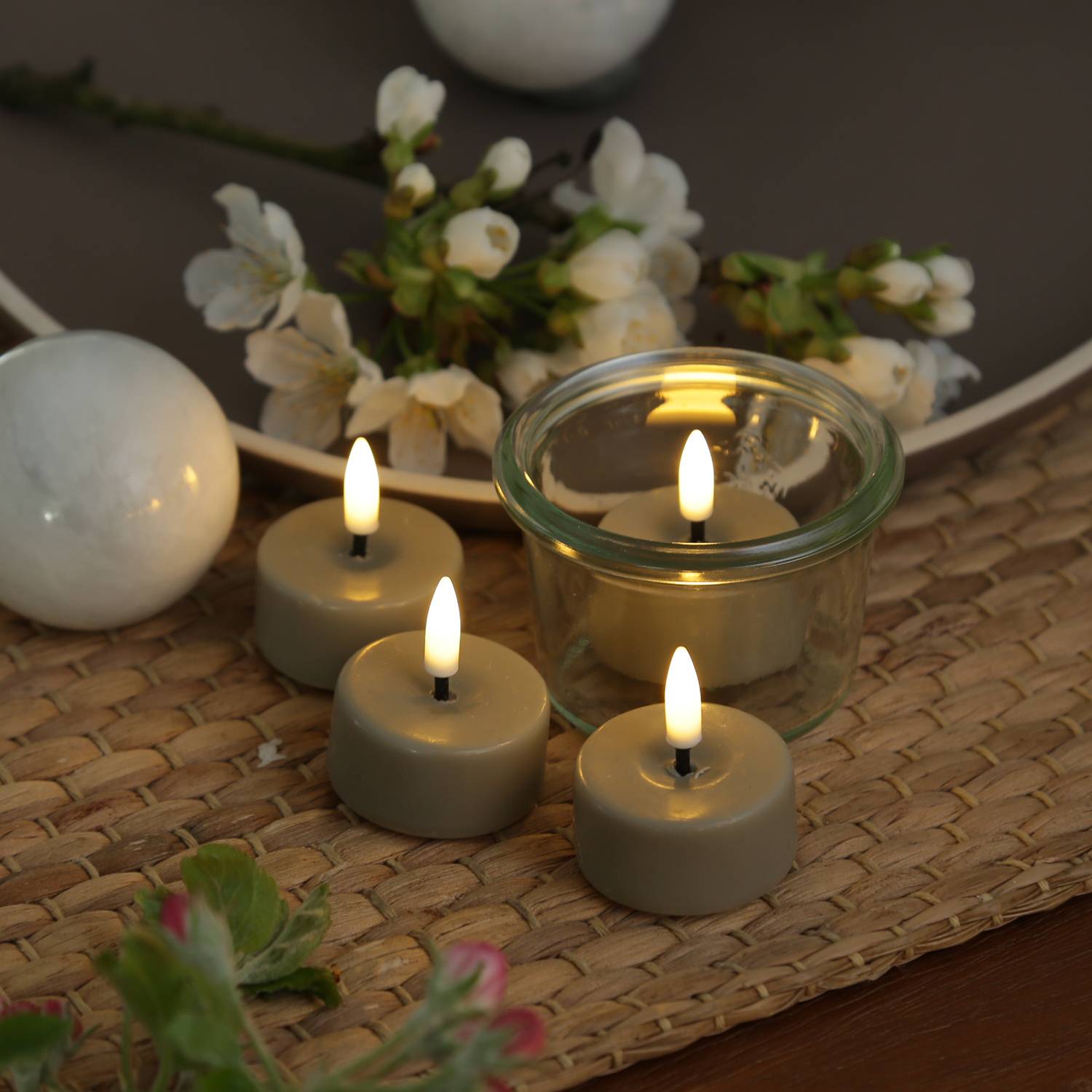 Vier angezündete Teelichter auf einer geflochtenen Matte, umgeben von weißen Blumen und Steinen, schaffen eine beruhigende, friedvolle Atmosphäre.