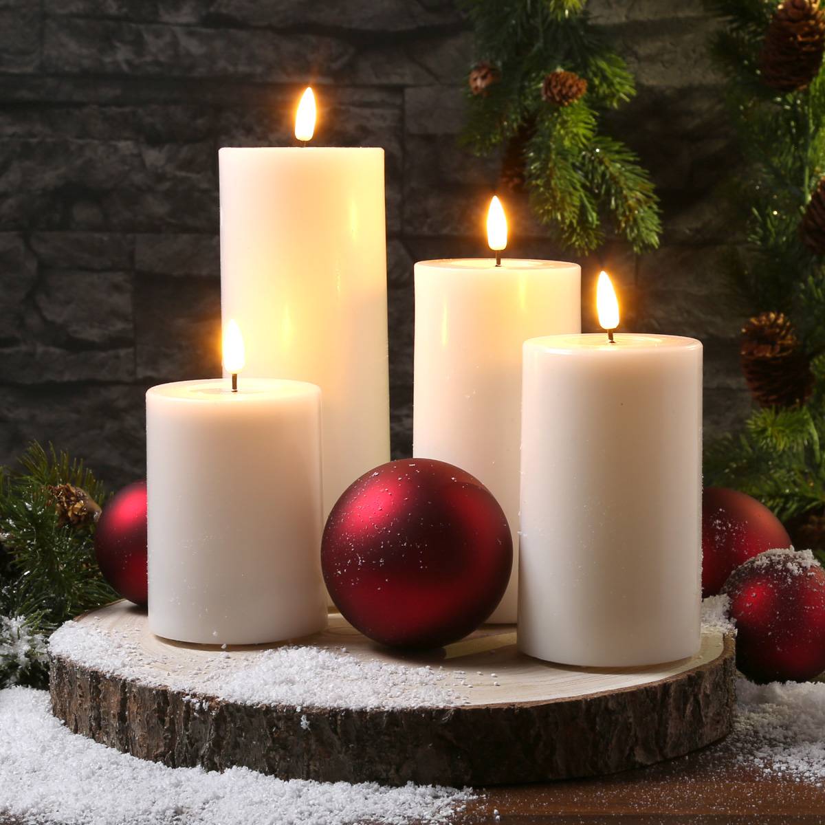 Vier brennende weiße Kerzen auf einem Holzbrett, umgeben von Zweigen immergrüner Nadelbäume und roten Weihnachtsschmuck, vor einer dunklen Ziegelwand.