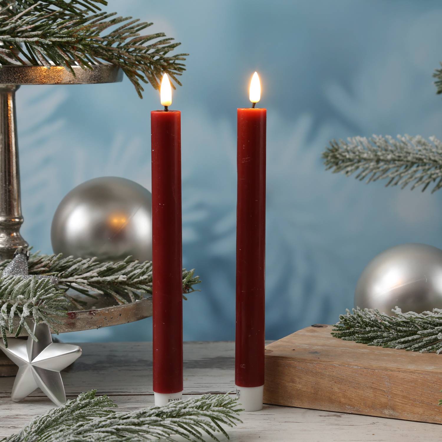Zwei rote Kerzen brennen auf einem festlich gedeckten Tisch mit Zweigen von Nadelbäumen und silbernen Ornamenten und schaffen eine gemütliche Feiertagsatmosphäre.