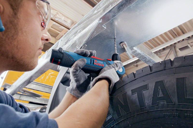 Eine Person benutzt einen Bosch-Akkuschrauber, um ein Metallblech unter einer Fahrzeughebebühne zu befestigen. Die Umgebung scheint eine Autoreparaturwerkstatt zu sein.