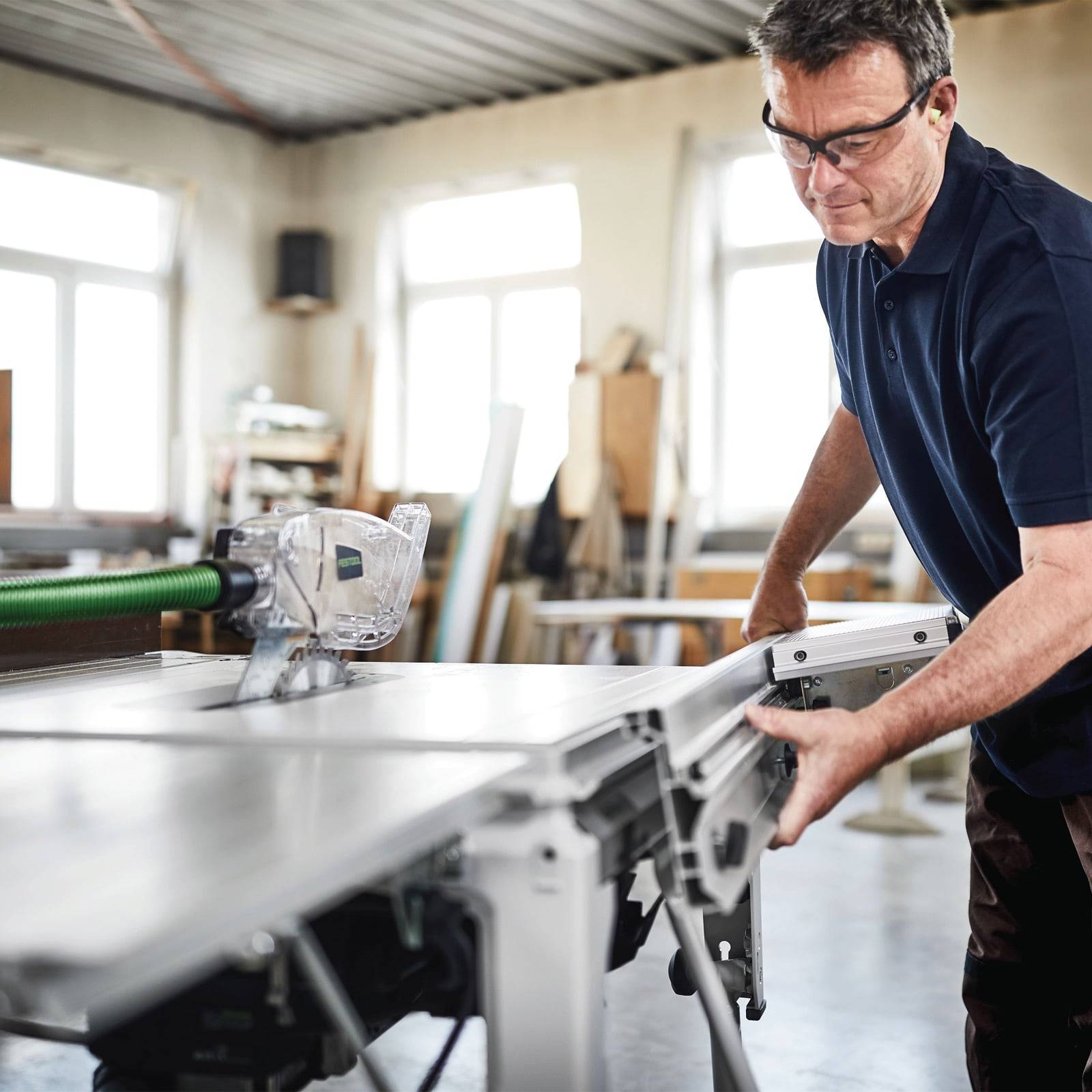 Ein Mann mit Schutzbrille lehnt sich über eine Kreissäge in einer Werkstatt und konzentriert sich auf präzise Arbeit. Sonnenlicht strömt durch große Fenster.