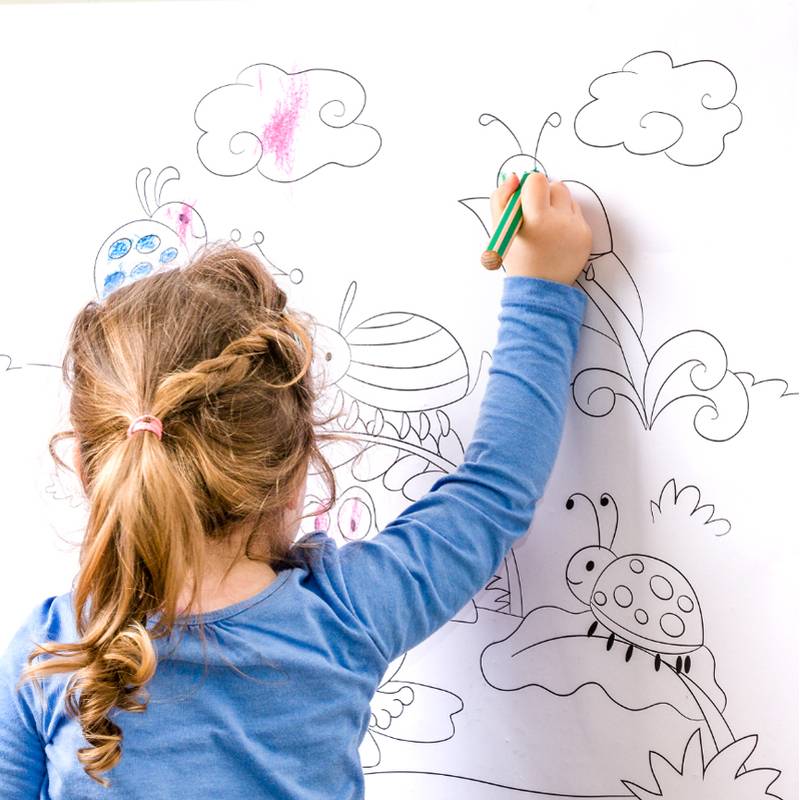 Ein junges Kind mit geflochtenen Haaren steht und zeichnet farbenfroh Marienkäfer und Wolken auf einer großen Whiteboard-Tafel und zeigt dabei Kreativität und Fantasie.