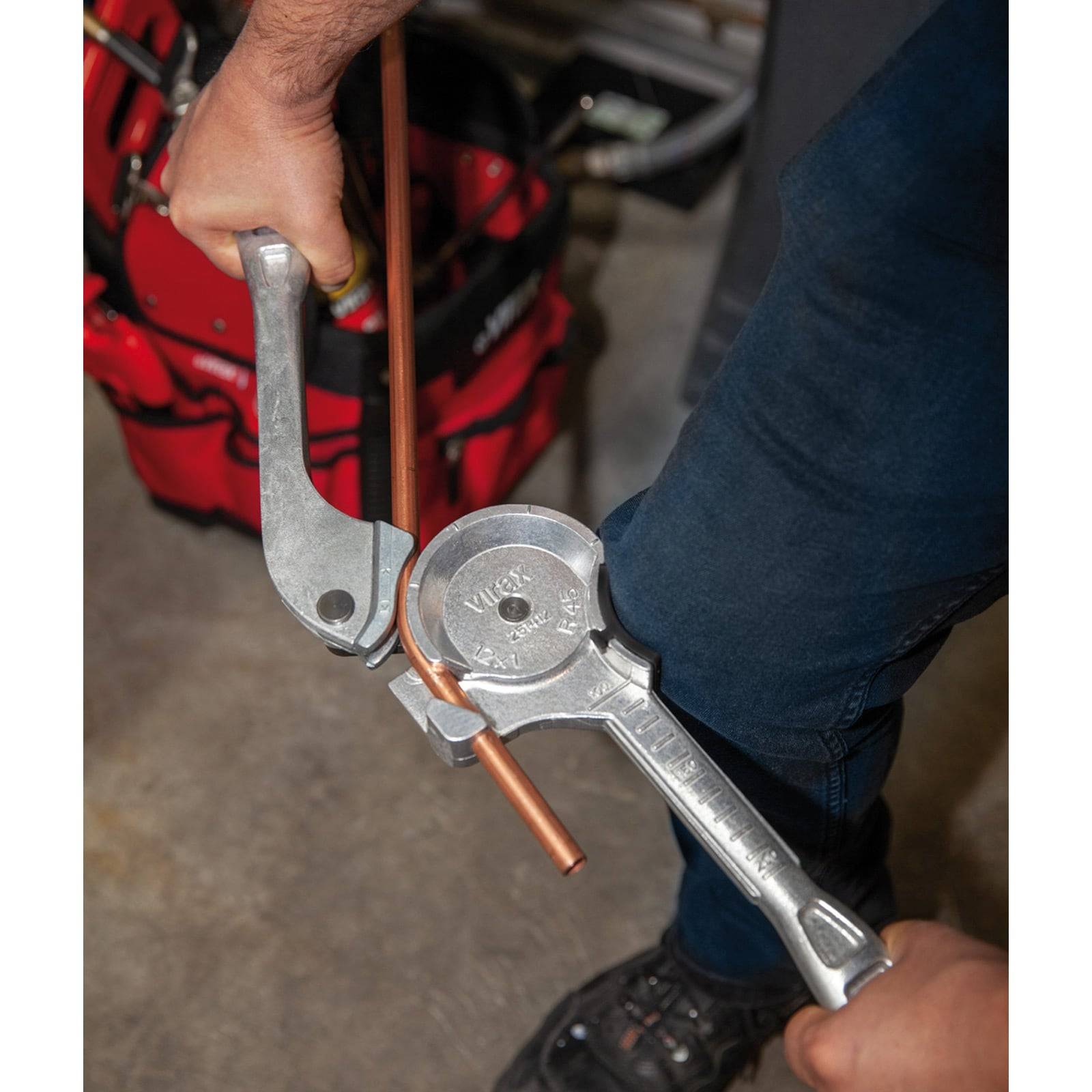 Eine Person benutzt einen Rohrbiegeapparat aus Metall, um ein Kupferrohr zu biegen, wobei eine rote Werkzeugtasche im Hintergrund sichtbar ist. Der Fokus liegt auf dem Biegeprozess.
