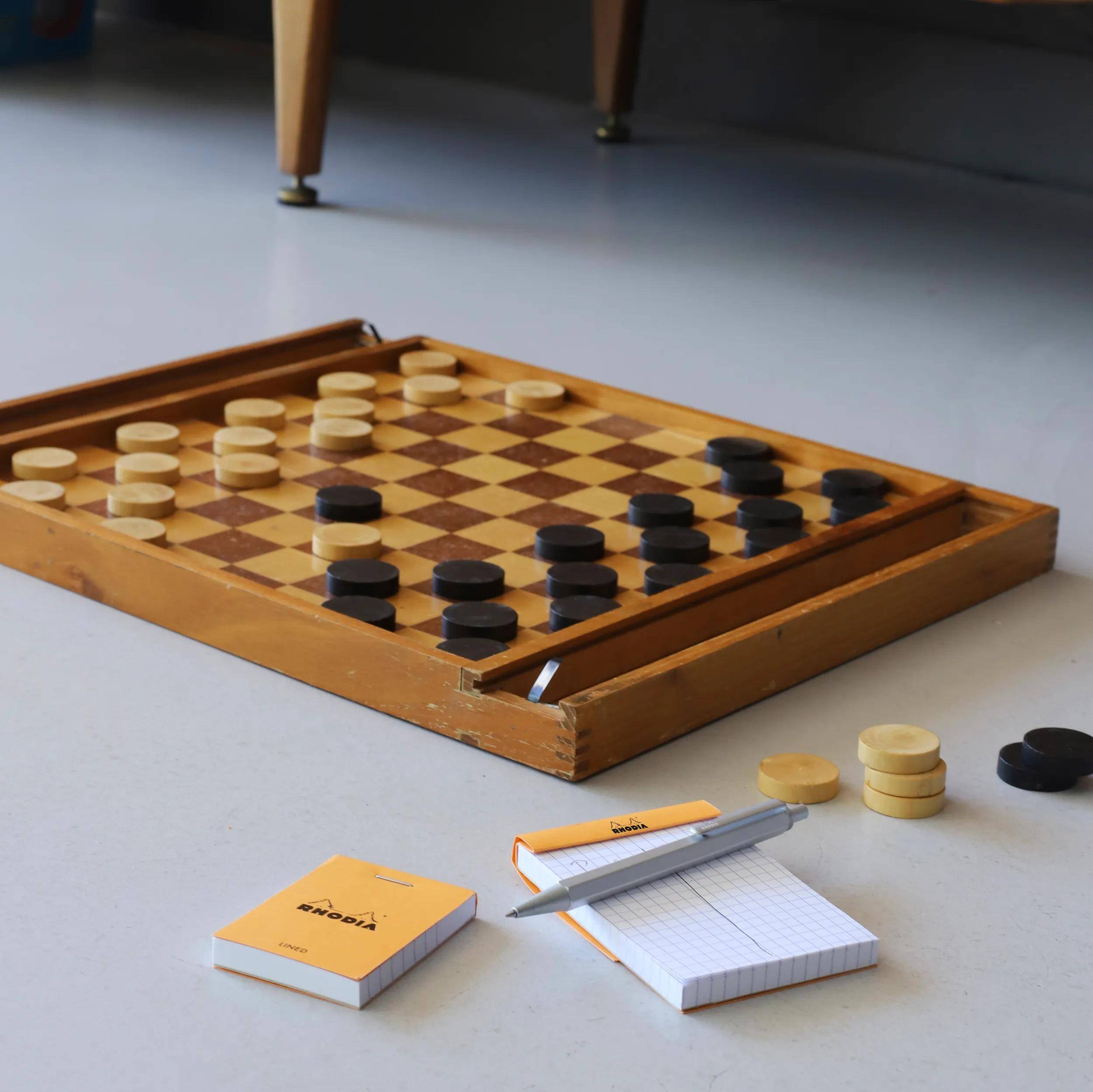 Ein Schachbrett aus Holz mit Spielsteinen im Spiel; verstreut herum sind ein Notizbuch, ein Stift und zusätzliche Spielsteine auf einem grauen Boden.