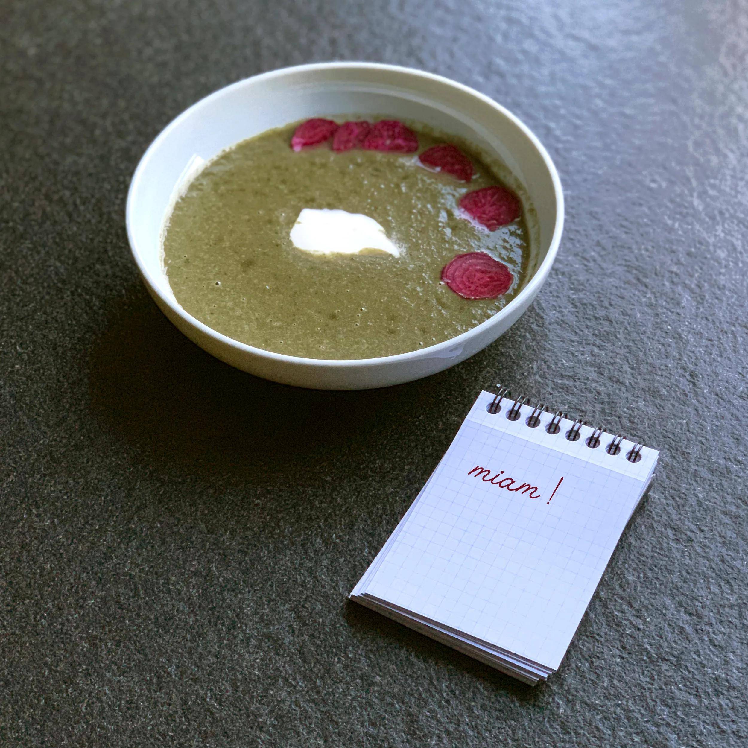 Eine Schüssel mit grüner Suppe, garniert mit Rübenscheiben auf einer dunklen Oberfläche, neben einem Notizblock mit dem Wort 