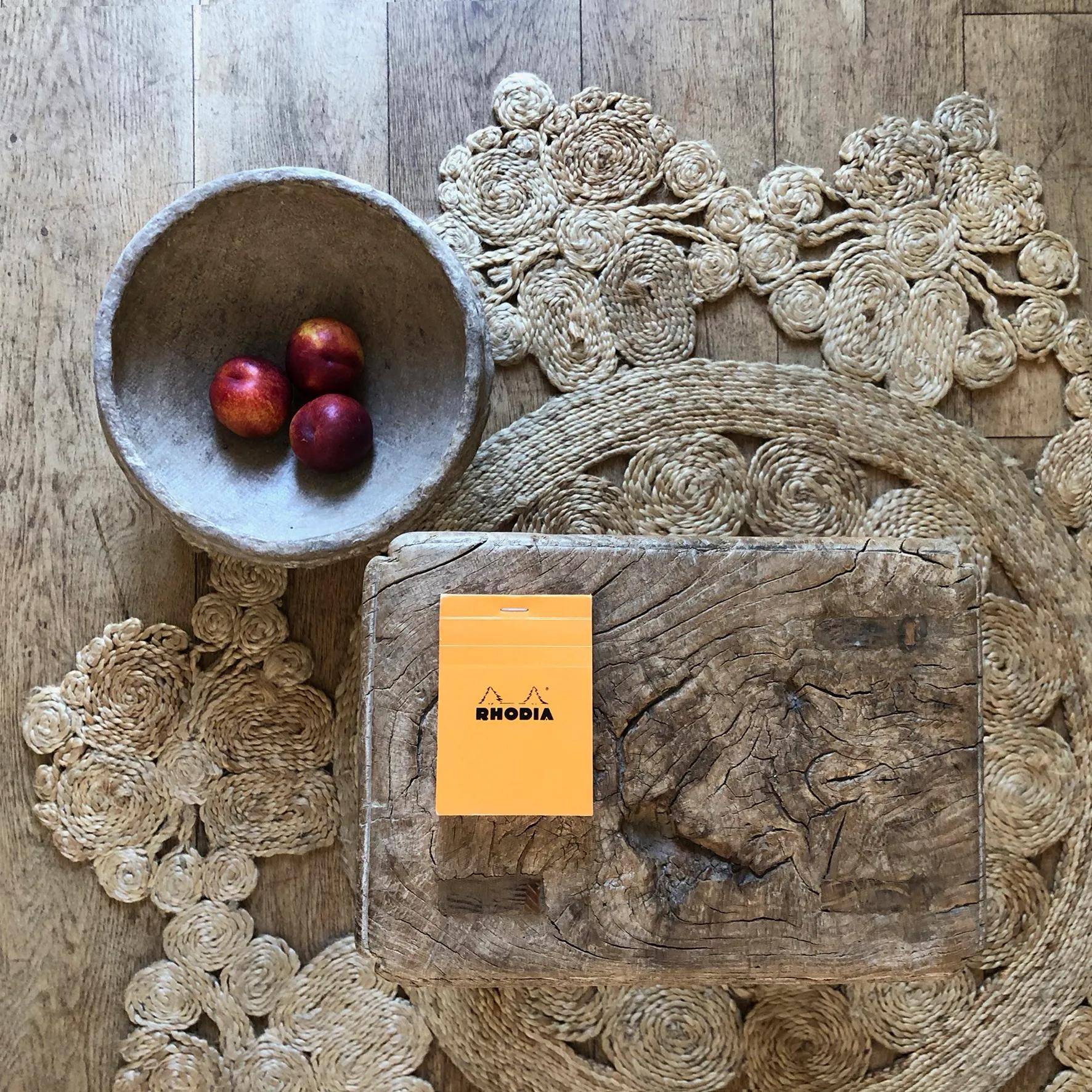 Ein kleines Notizbuch mit orangefarbenem Umschlag mit der Aufschrift Rhodia liegt auf einem rustikalen Holztisch. In der Nähe befindet sich ein Korb mit drei roten Äpfeln.