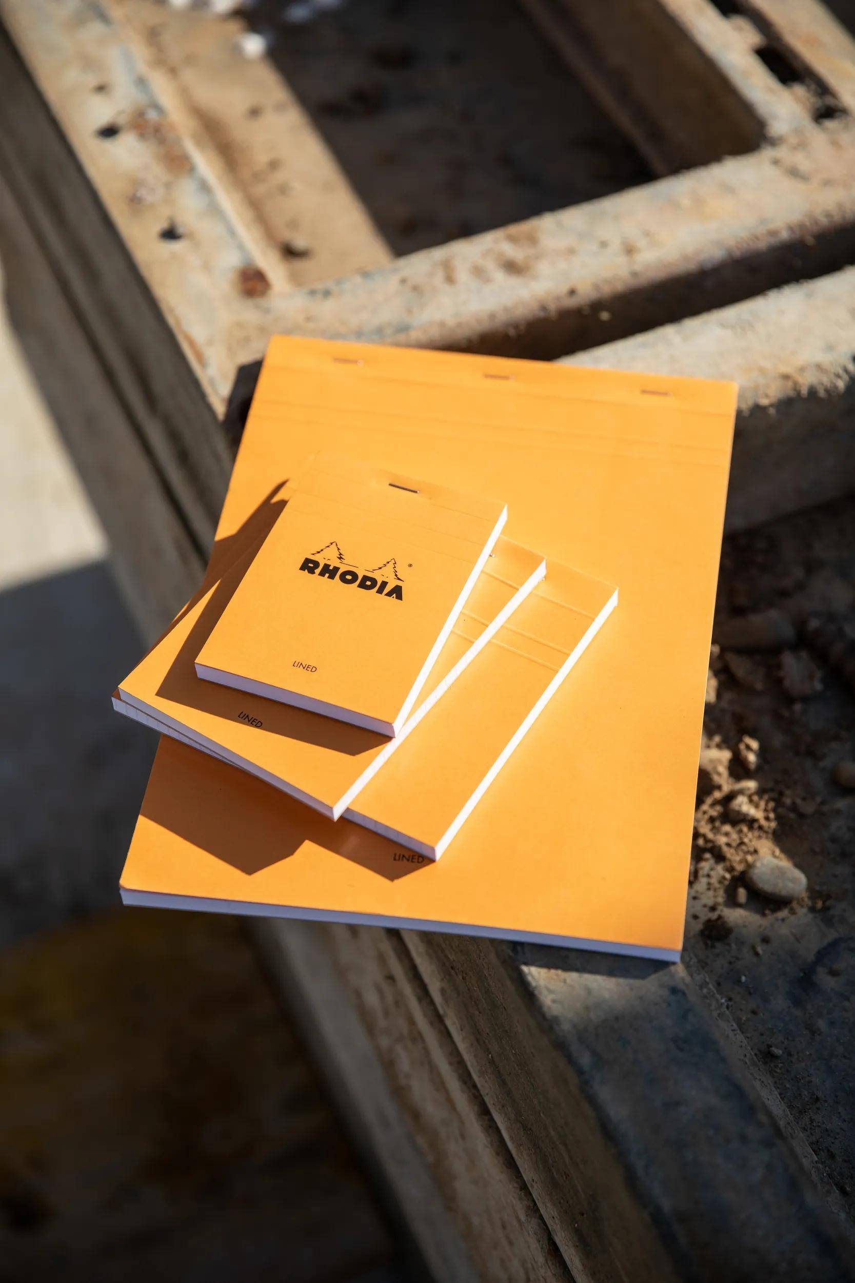 „Drei orangefarbene Rhodia-Notizblöcke in verschiedenen Größen auf einer strukturierten Oberfläche, mit Sonnenlicht, das Schatten wirft und Außennutzung andeutet.