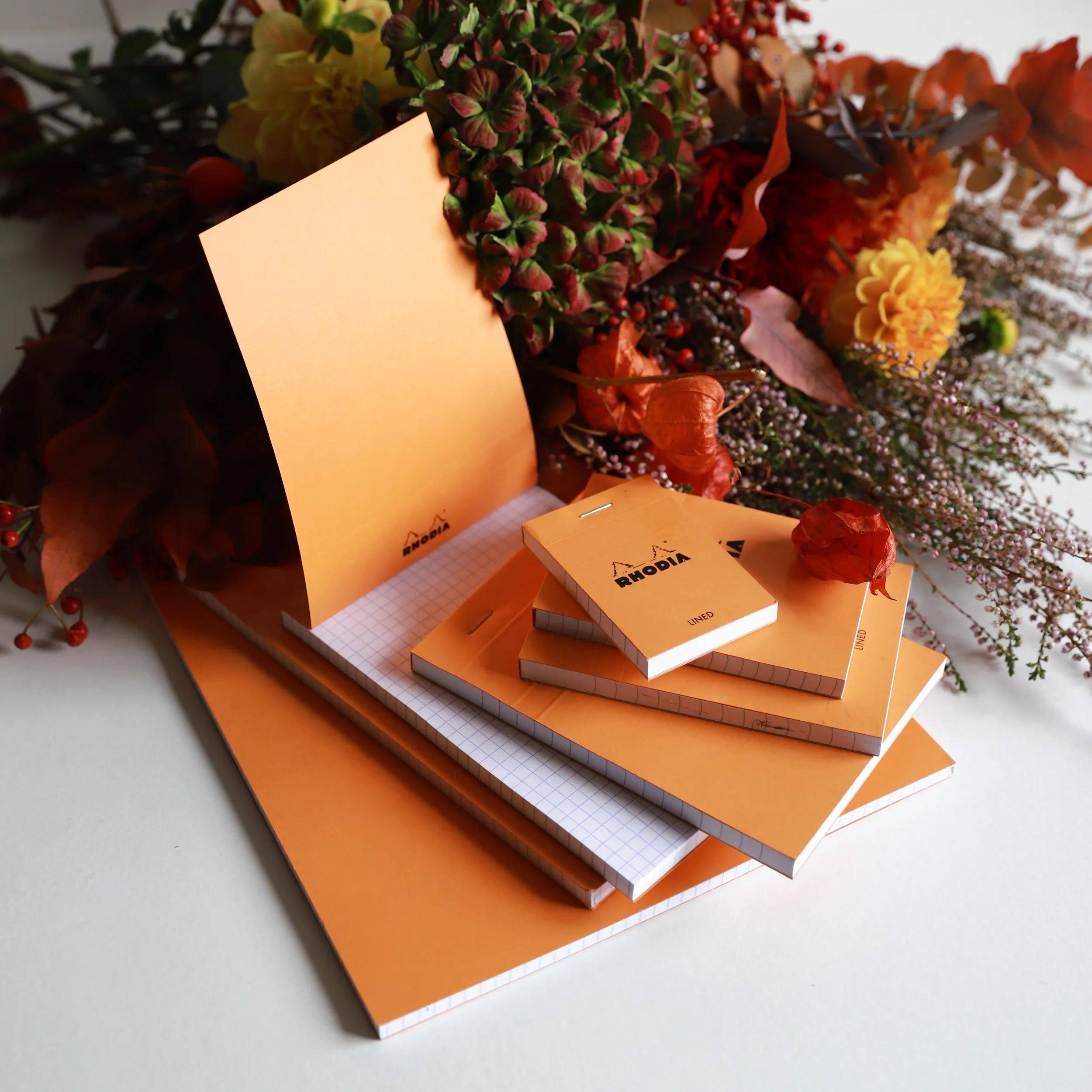 Ein Stapel orangefarbenes Schreibwaren und Notizblocke sitzt auf einer weißen Oberfläche, umgeben von lebendigen Herbstblumen und Blättern.