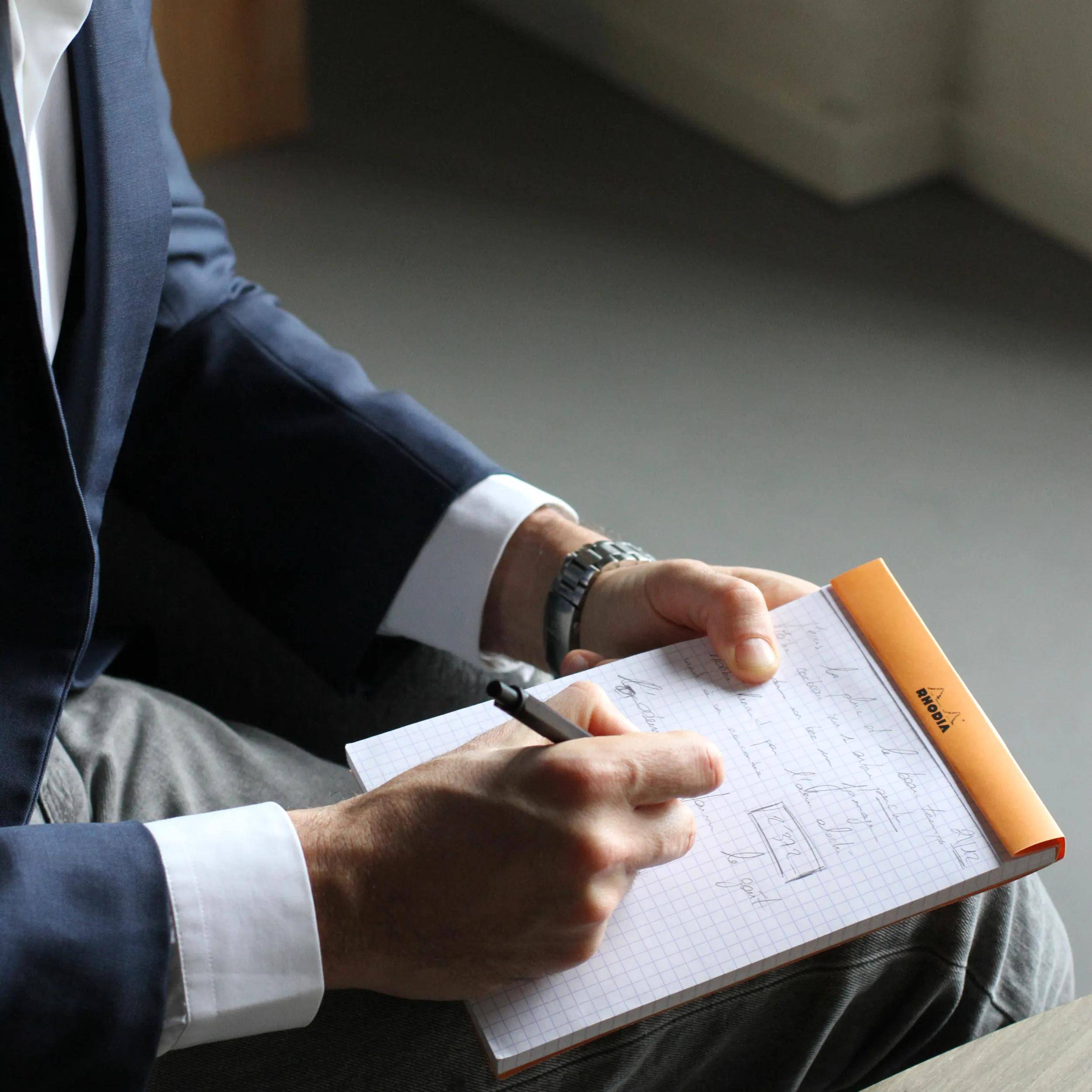 Eine Person in einem Anzug schreibt Notizen auf einem Klemmbrett mit Millimeterpapier. Der Fokus liegt auf ihren Händen und dem Stift, was aktives Engagement anzeigt.