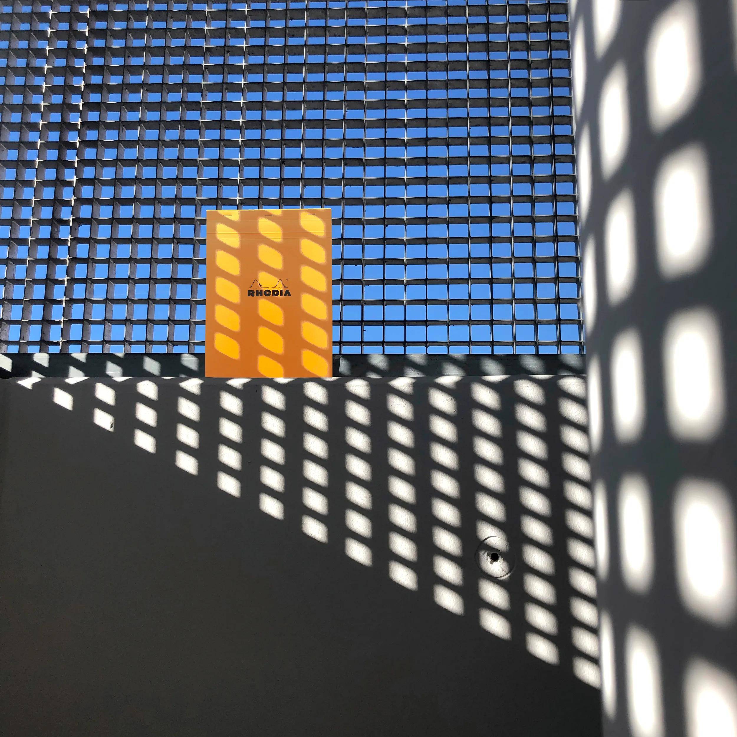 Ein leuchtend oranges Notizbuch steht vor einem gittermustrigen Fenster und wirft komplexe Schatten auf eine graue Oberfläche, wobei ein blauer Himmel sichtbar ist.