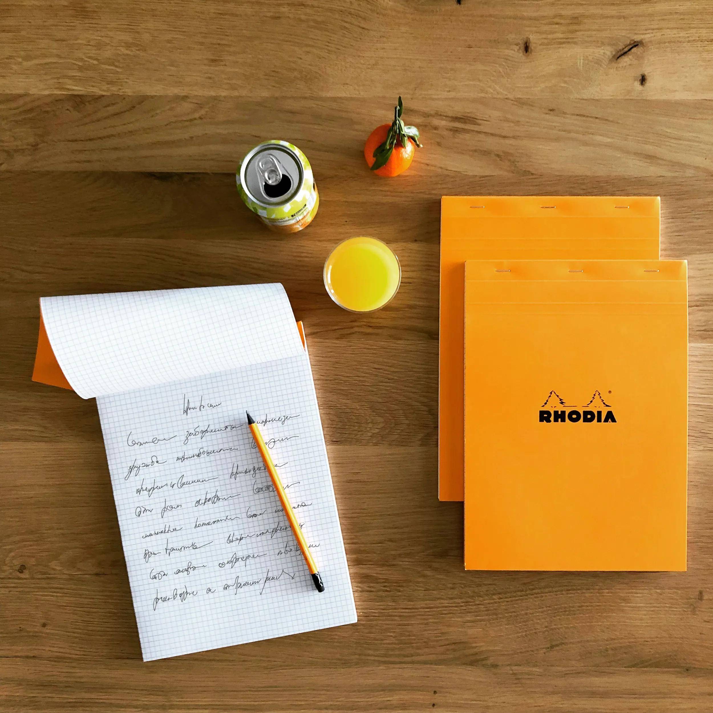 Draufsicht auf einen Schreibtisch mit einer handschriftlichen Notiz auf Millimeterpapier, einem Bleistift, orangefarbenen Rhodia-Notizbüchern, einer Dose Limonade, einer kleinen orangefarbenen Frucht und einem Glas Saft.