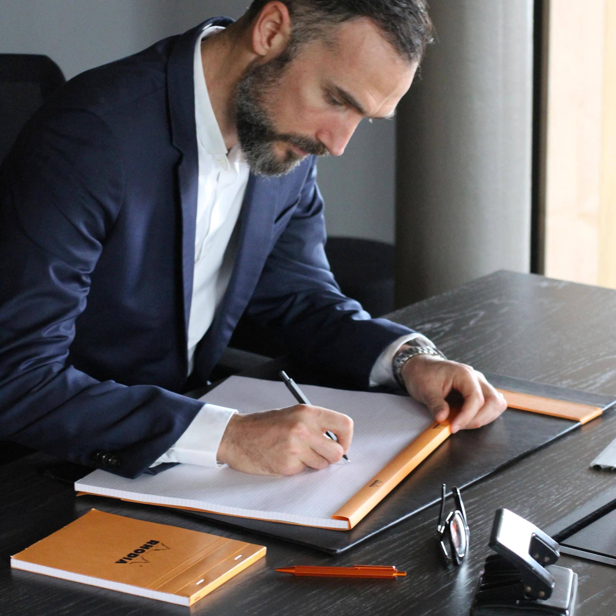 Ein Mann in einem Anzug sitzt an einem Schreibtisch und schreibt mit einem Stift auf Papier. Der Schreibtisch hat einen Laptop, einen Notizblock und eine Brille. Die Umgebung ist professionell.