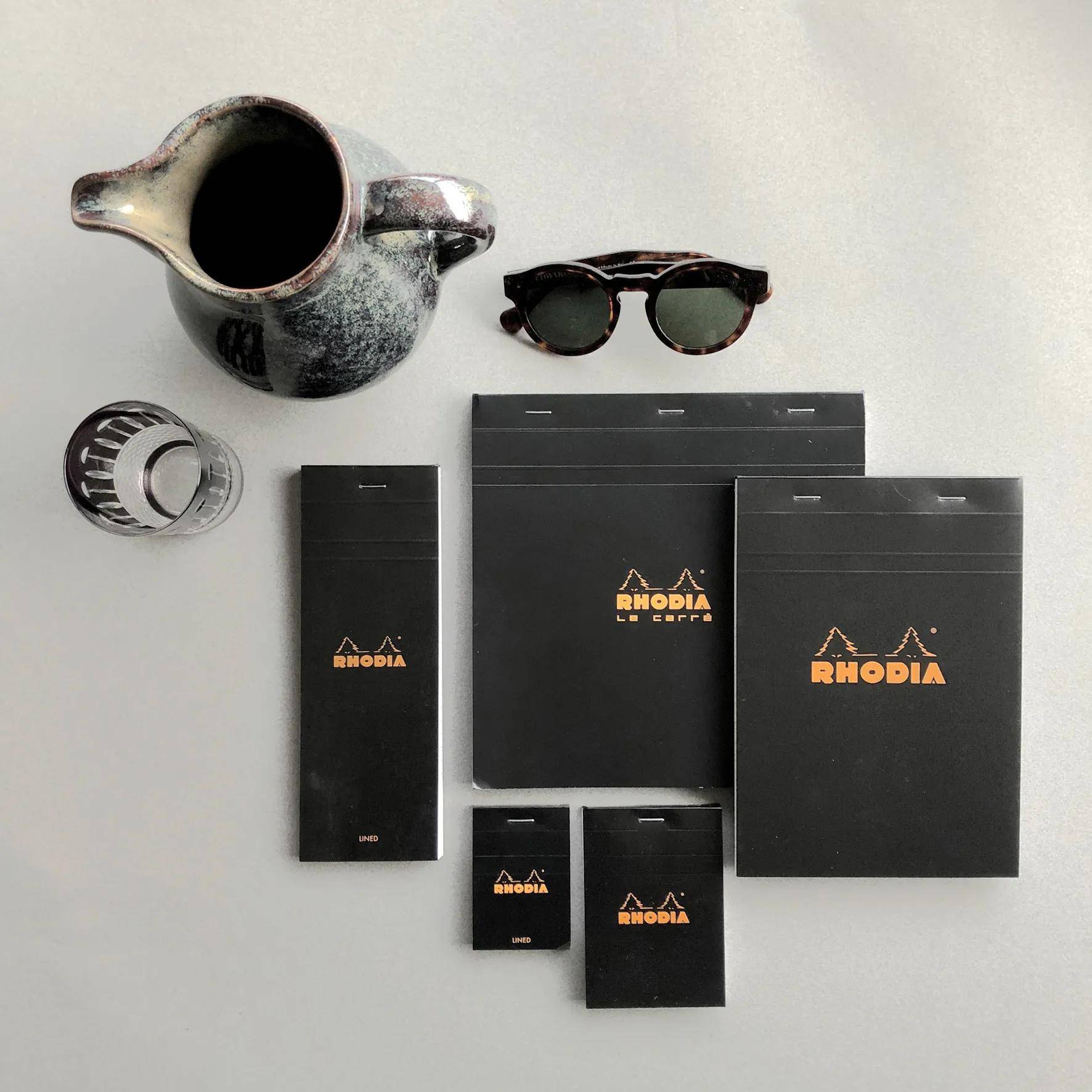 Eine Sammlung von Rhodia-Notizbüchern in verschiedenen Größen, die auf einem Tisch neben einem Keramikkrug, einem Glas und einer Sonnenbrille angeordnet sind.
