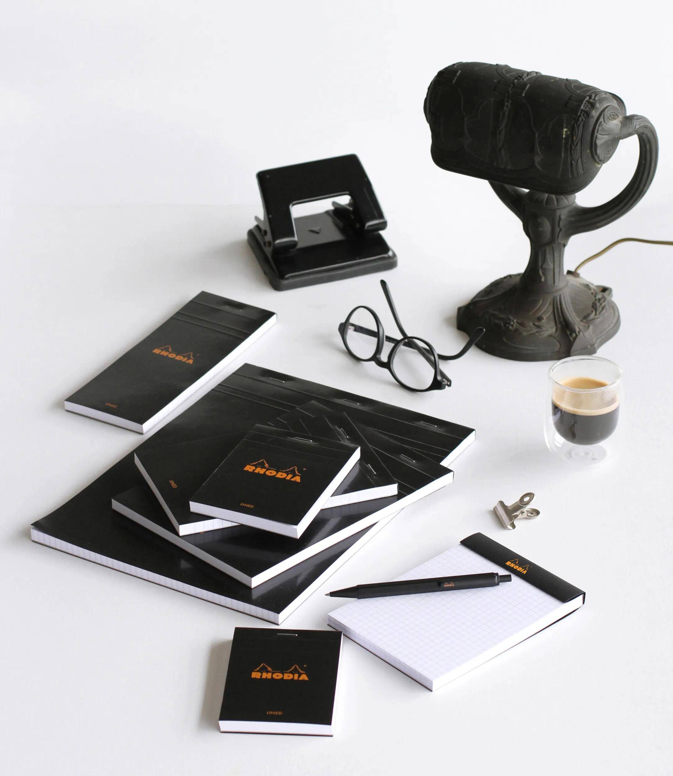 Büroschreibtisch mit Rhodia-Notizblöcken, einer Vintage-Lampe, einer Brille, einem Locher, einer Kaffeetasse, einer Büroklammer und einem Stift, was auf einen Arbeitsplatz hindeutet.