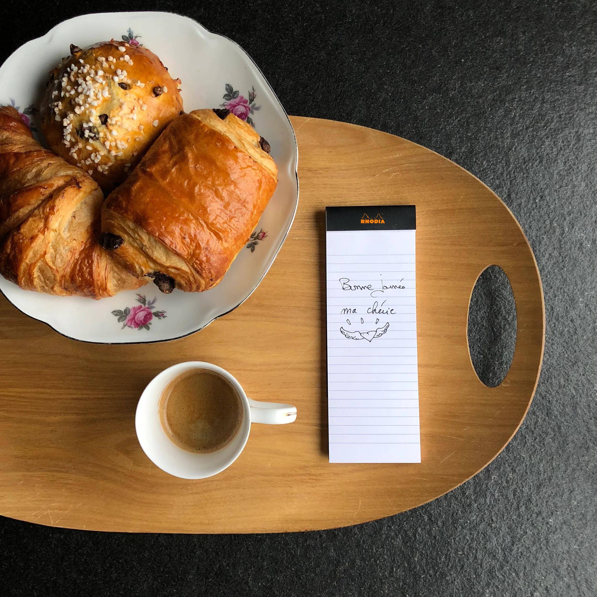 Ein hölzernes Tablett mit einem Croissant, Pain au Chocolat und einem mit Zucker bestrichenen Gebäck. Eine Tasse Espresso und ein Notizblock mit einer Notiz 