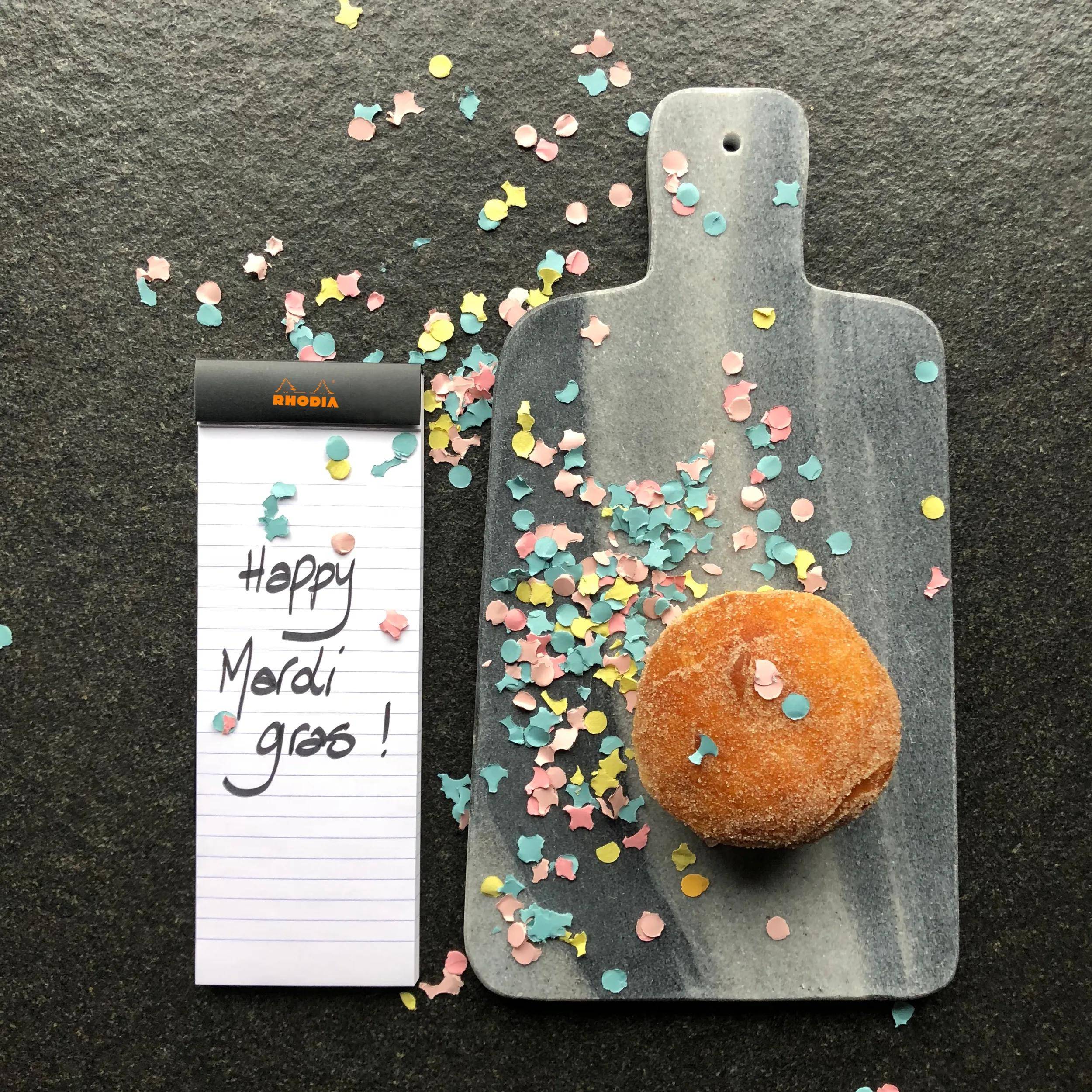 Ein Schneidebrett mit einem gepuderten Donut daneben, neben einem Notizblock mit der Aufschrift „Happy Mardi Gras!