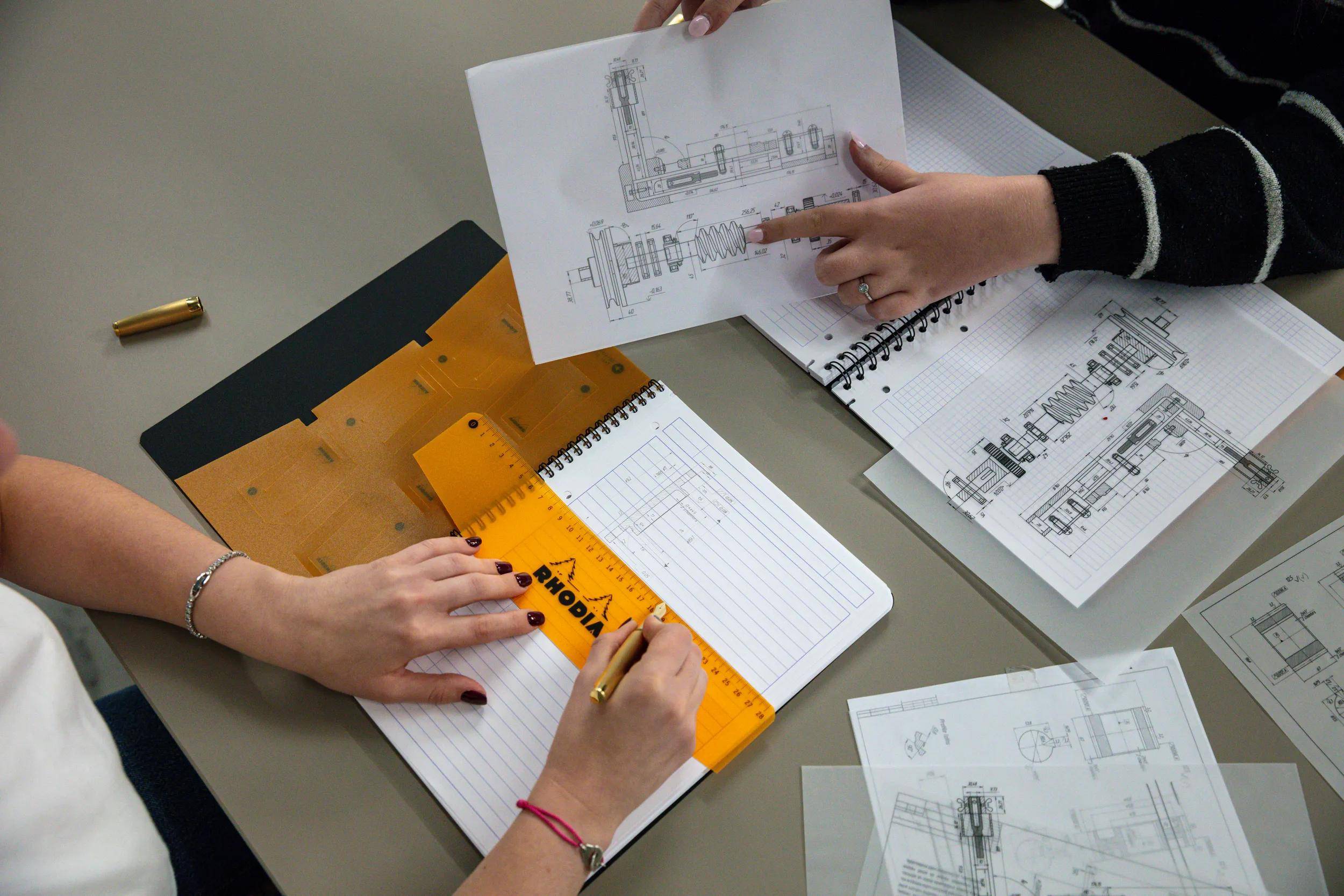 Zwei Personen arbeiten zusammen an technischen Bauplänen und nutzen dabei Stahllineal und Zeichenwerkzeuge, wobei sie sich auf Genauigkeit und Designdetails konzentrieren.