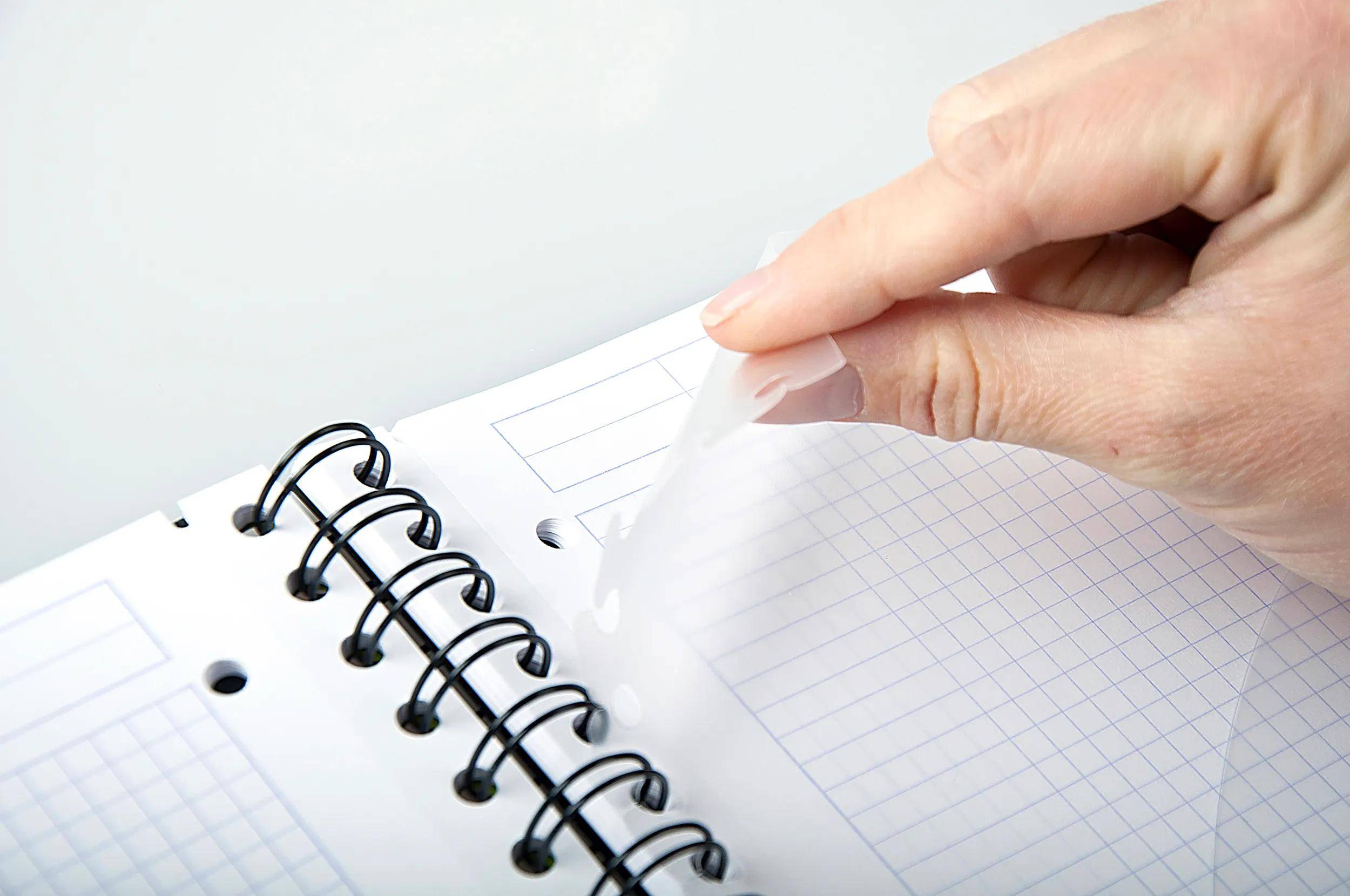 Eine Person reißt eine Seite aus einem spiralgebundenen Notizbuch heraus, das Gitter- und linierte Abschnitte zeigt und auf Planung oder Notizen hindeutet.