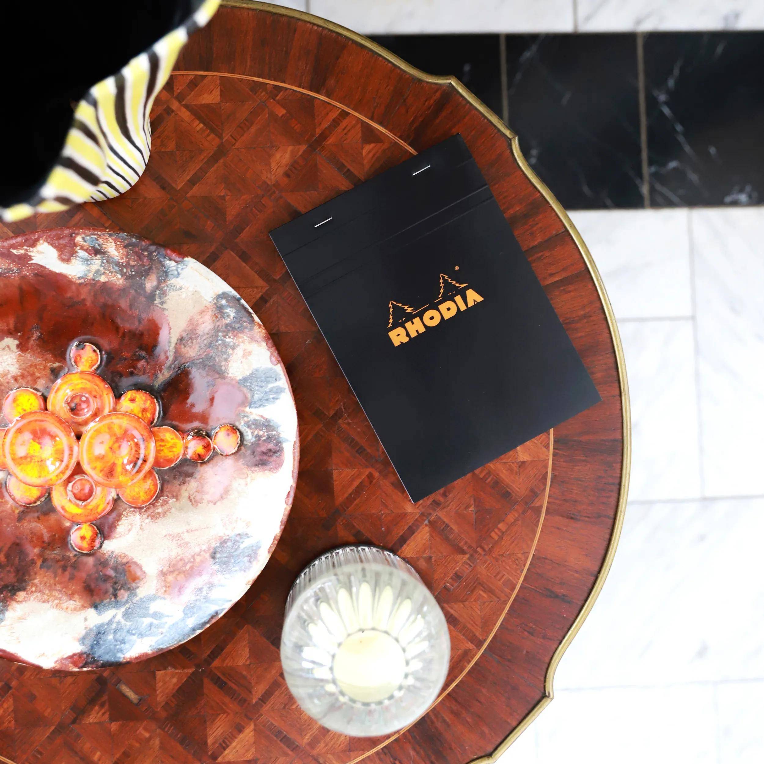 Ein 'Rhodia'-Notizblock liegt auf einem runden Holztisch mit einer dekorativen Schale gefüllt mit orangefarbenen Kugeln und einer brennenden Kerze, von oben betrachtet.