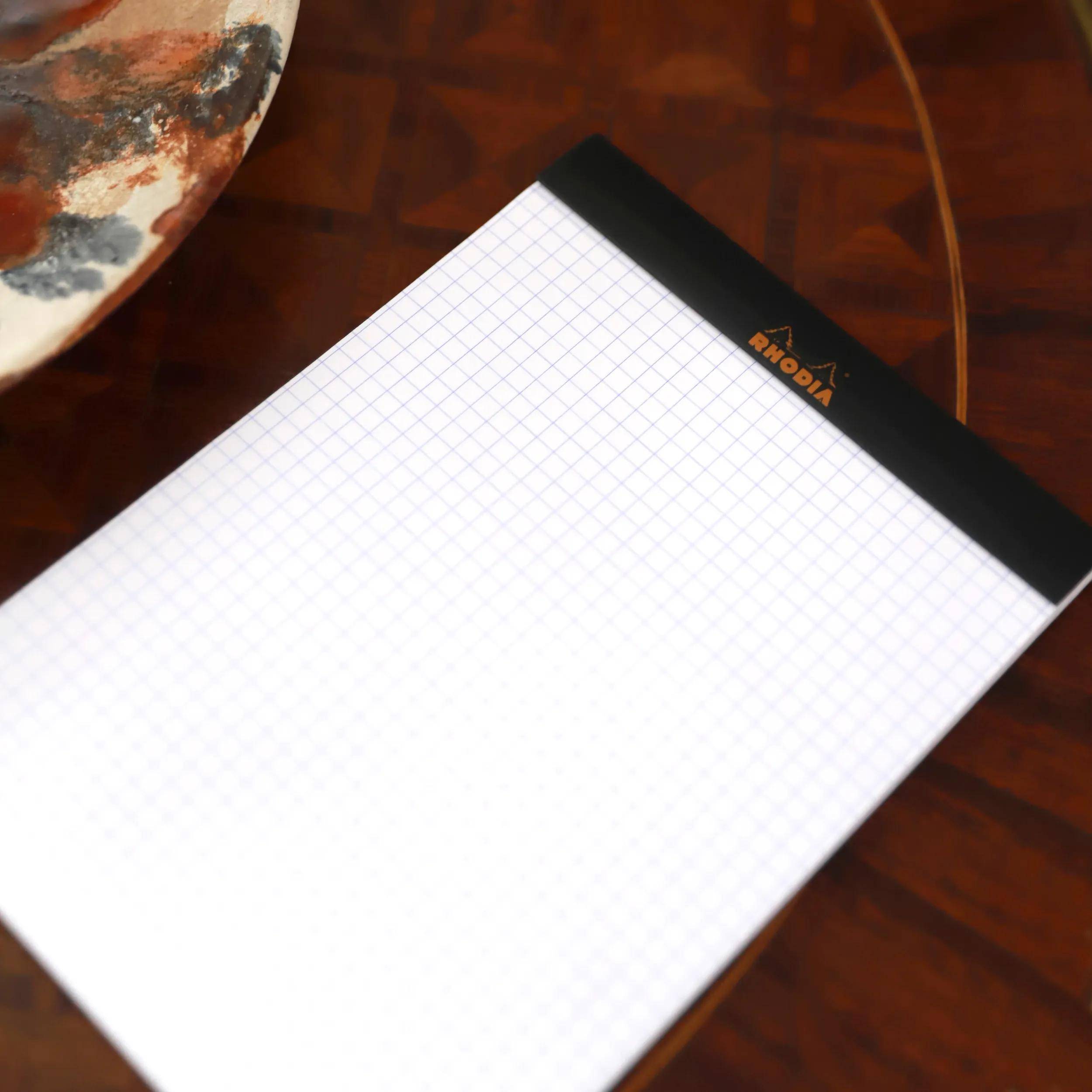 Karierter leerer Notizblock mit schwarzer Obenbindung und Rhodia-Logo auf einem Holztisch.