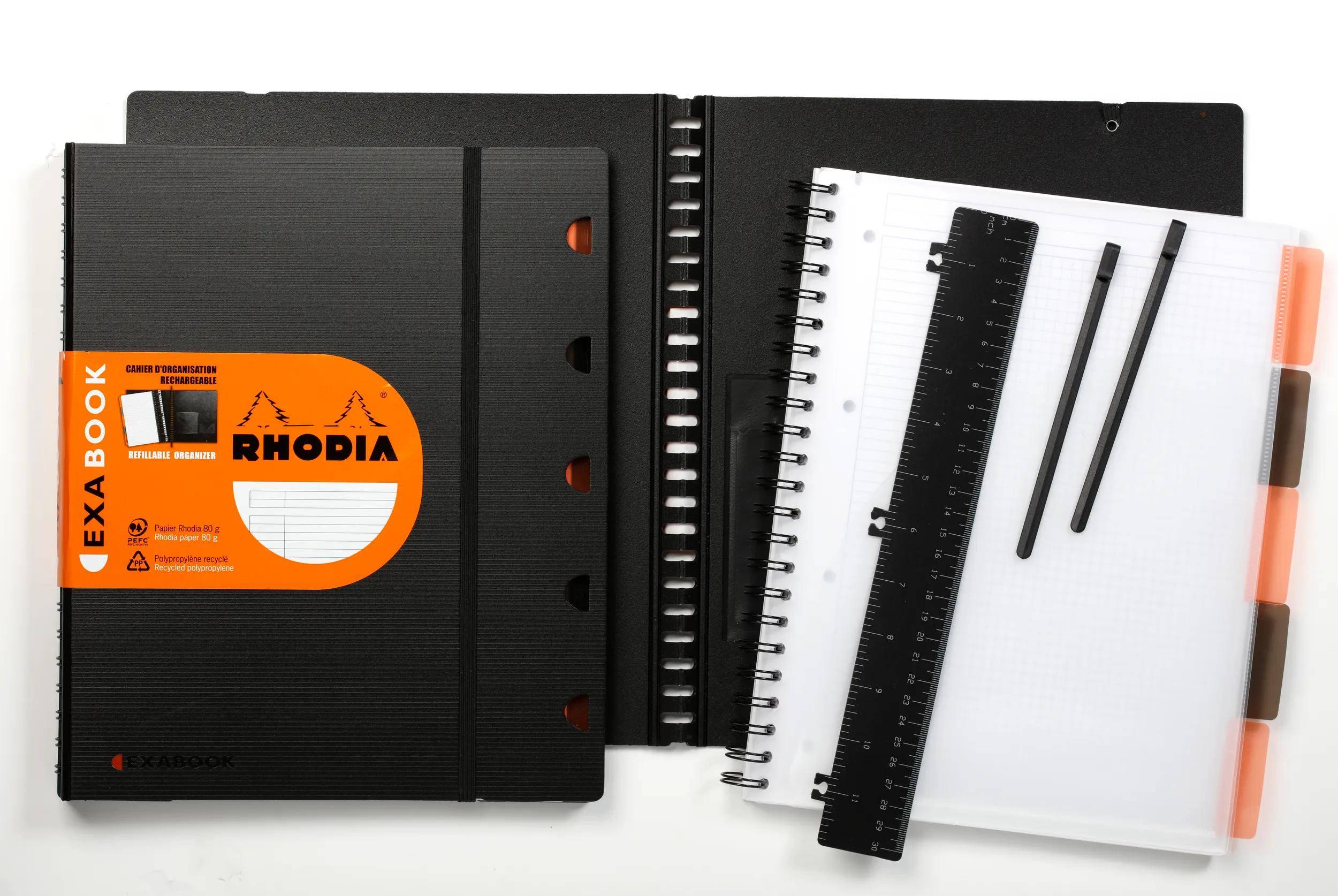 Ein 'RHODIA' Spiralblock in Schwarz und Orange liegt offen mit linierten Seiten, einem Lineal und Bleistiften auf einer weißen Oberfläche.