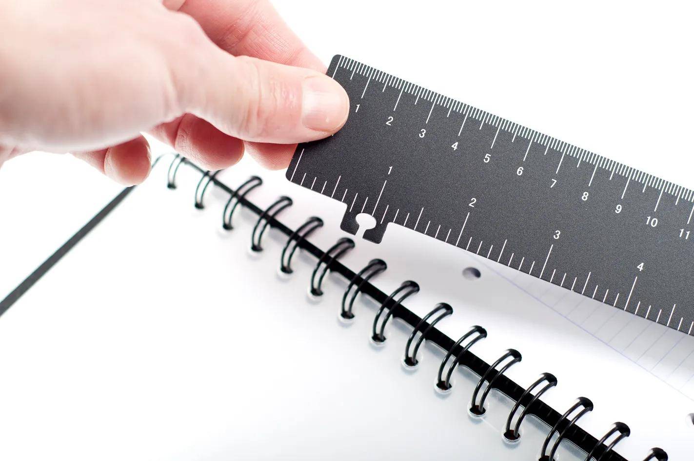 Eine Hand hält ein schwarzes Lineal über ein spiralgebundenes Notizbuch und zeigt eine genaue Messung des Abstands zwischen den Spiralringen.