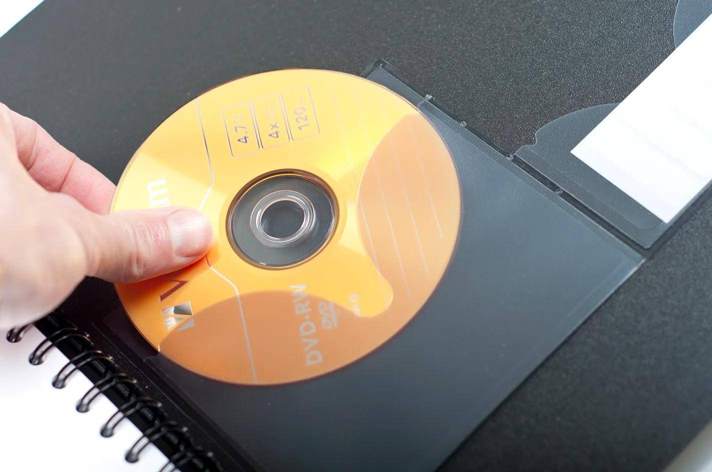 Eine Hand legt eine DVD-Scheibe in eine notizbuchähnliche Hülle, daneben liegt ein spiralgebundenes Notizbuch.