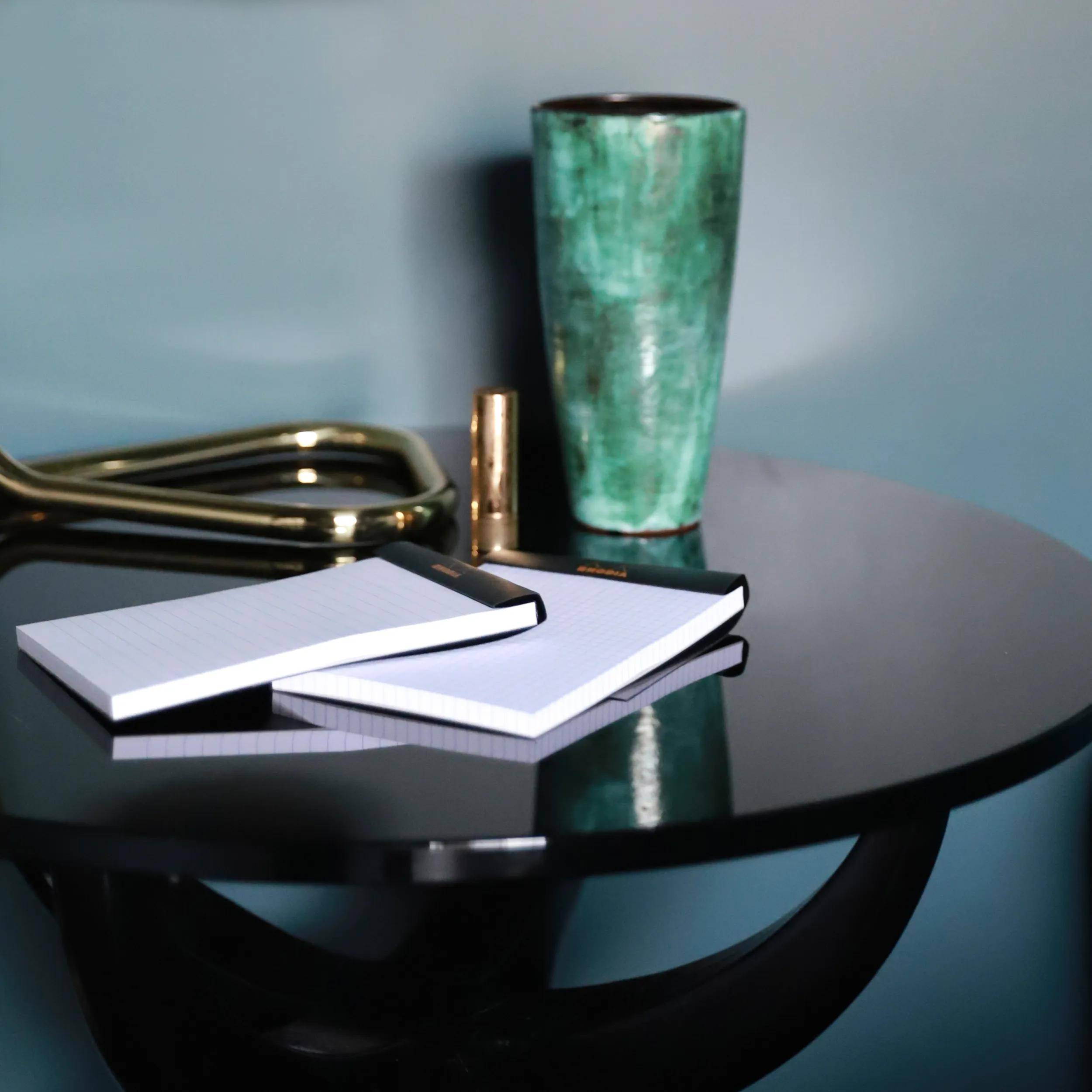 Ein schwarzer runder Tisch mit einer grünen Keramikvase, goldenen Scheren und zwei gestapelten Notizblöcken. Die Szene deutet auf eine Arbeitsplatzeinrichtung hin.