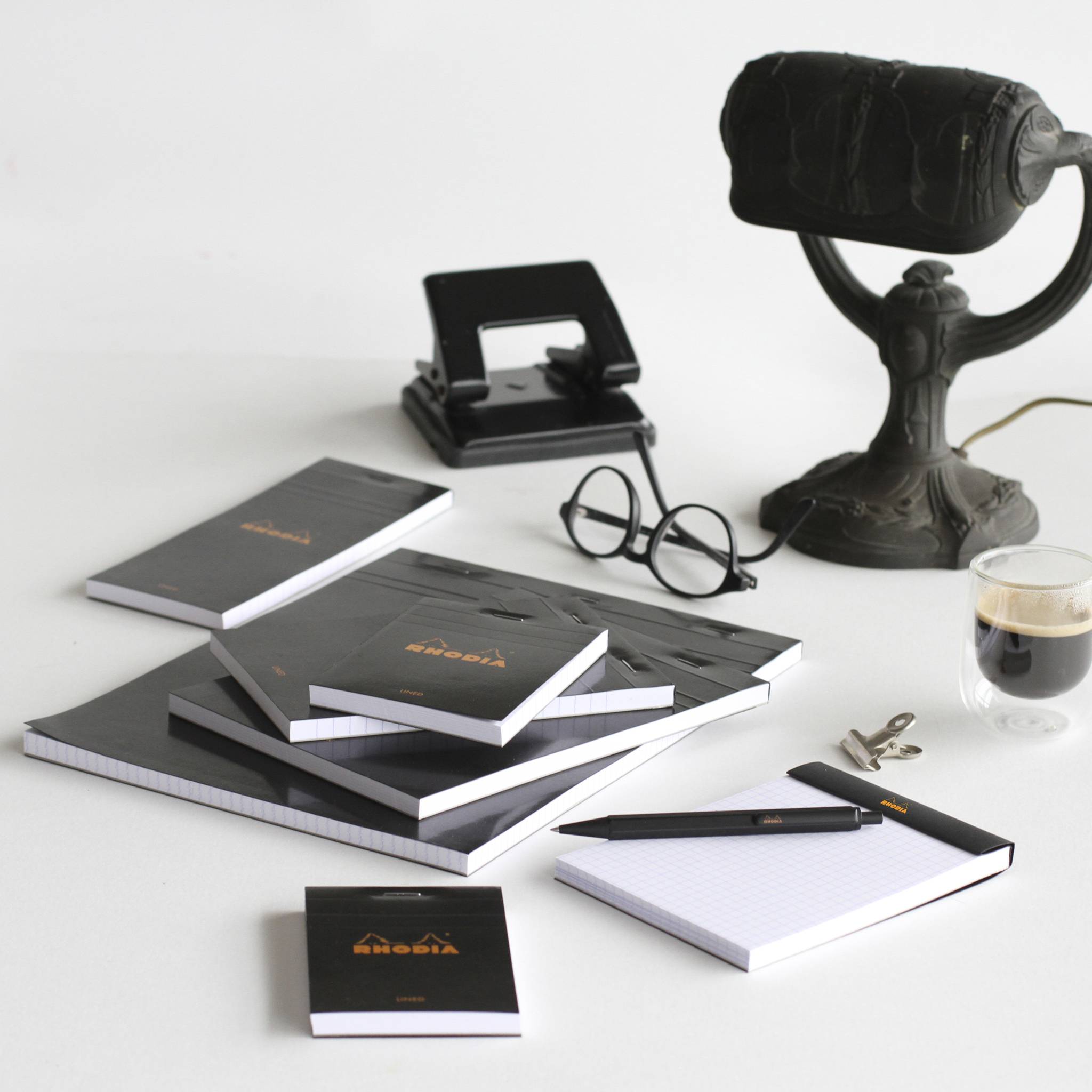Ein Stillleben auf einem Schreibtisch mit gestapelten schwarz-weißen Notizbüchern, einer Brille, einem Hefter, einem Stift, Schlüsseln, einer Kaffeetasse und einer Lampe im Vintage-Stil auf einer weißen Oberfläche.