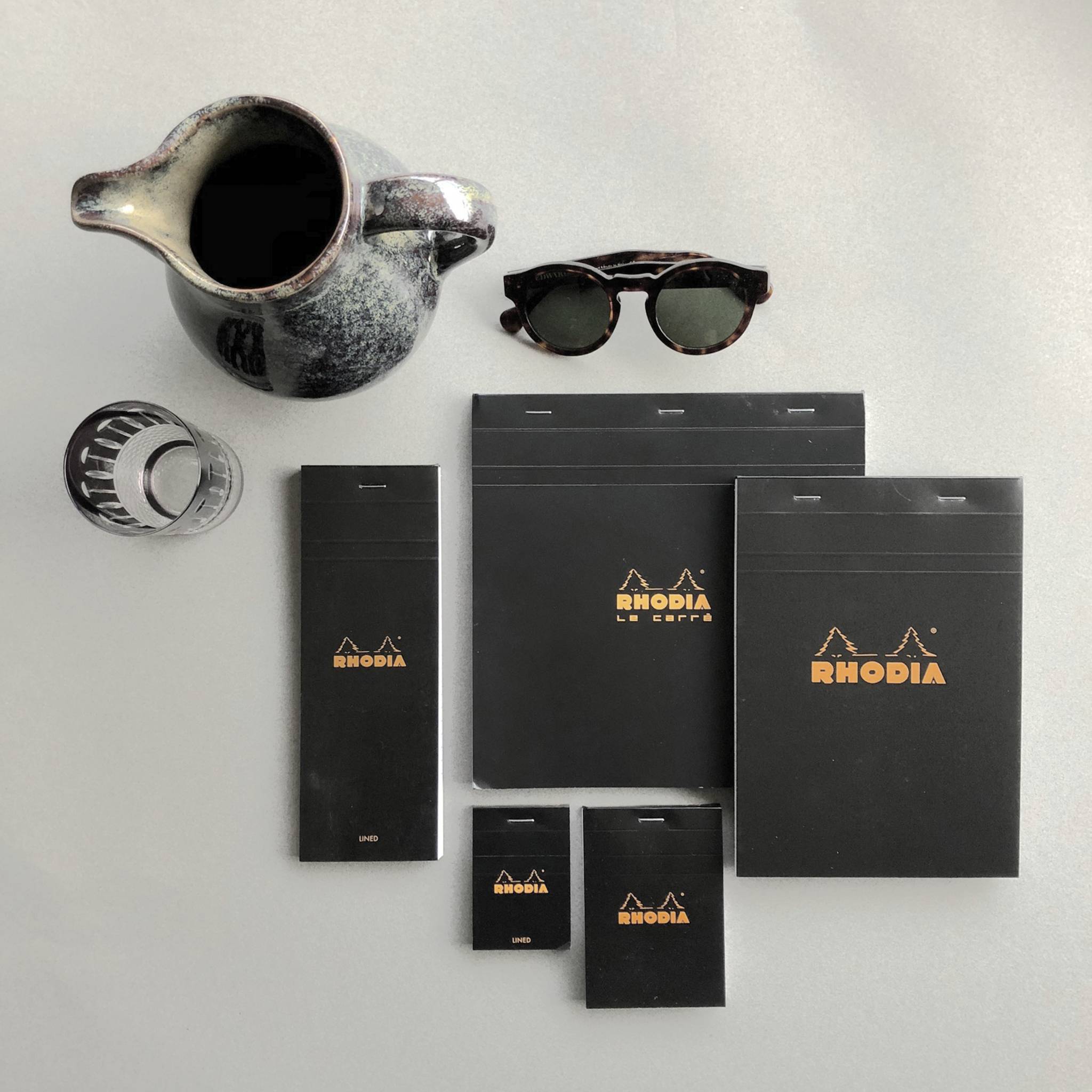 Verschiedene Rhodia-Schreibwaren und eine Sonnenbrille auf einer hellgrauen Oberfläche angeordnet, einschließlich Notizbücher, Blöcke und einer Metallkanne.