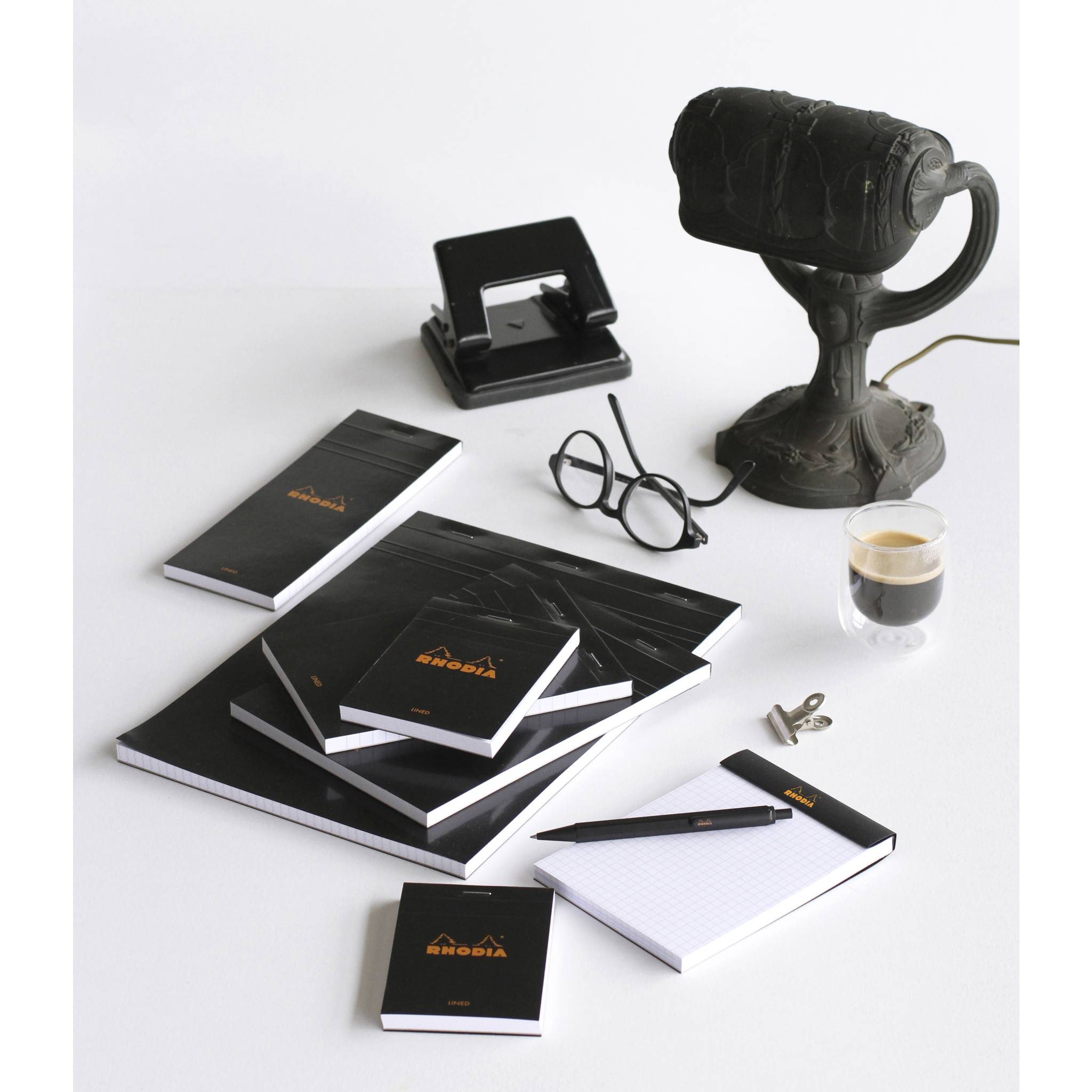 Markengebundene Büroschreibtischobjekte auf weißem Hintergrund: Notizbücher, ein Hefter, eine Brille, ein Stift, eine Kaffeetasse und eine Vintage-Schreibtischlampe.