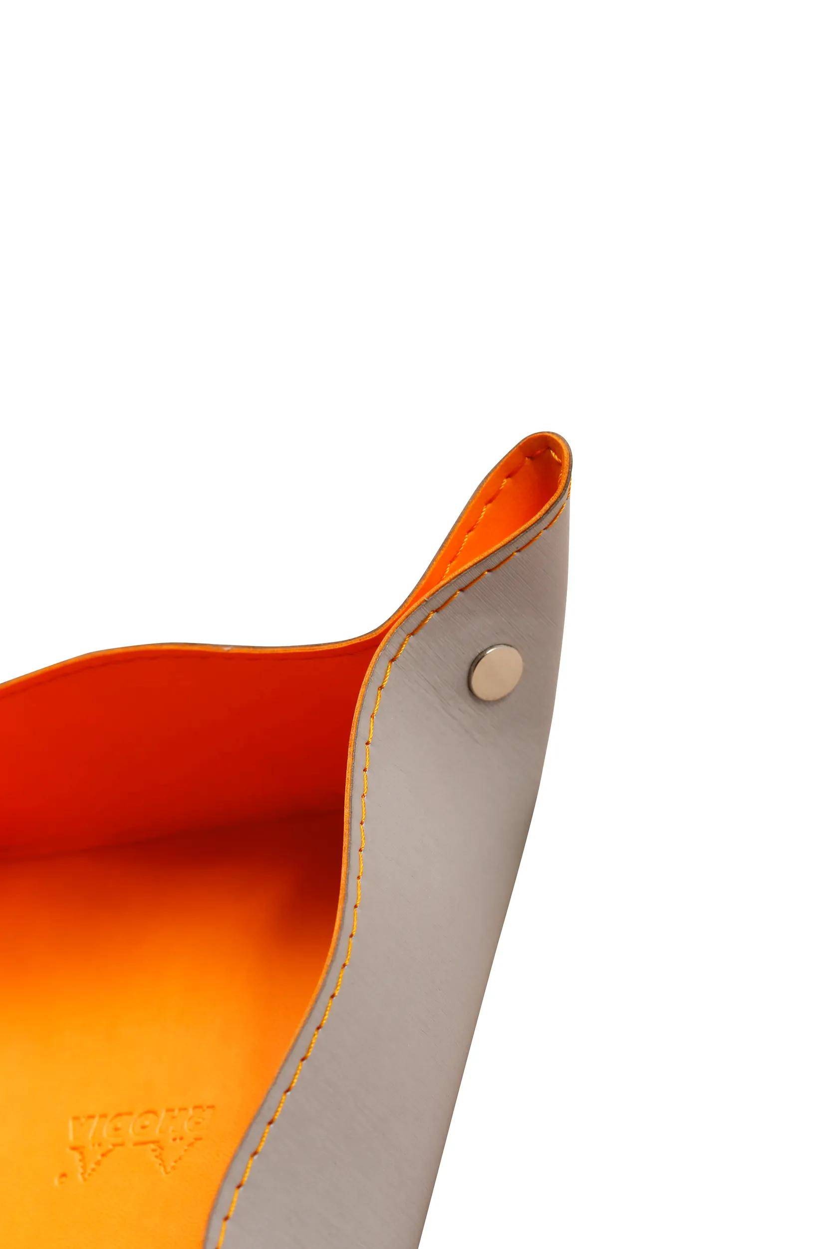 Ein Nahaufnahmebild einer orange-grauen Ledertasche, das ihre gebogene Kante und das silberfarbene Knopfdetail zeigt.