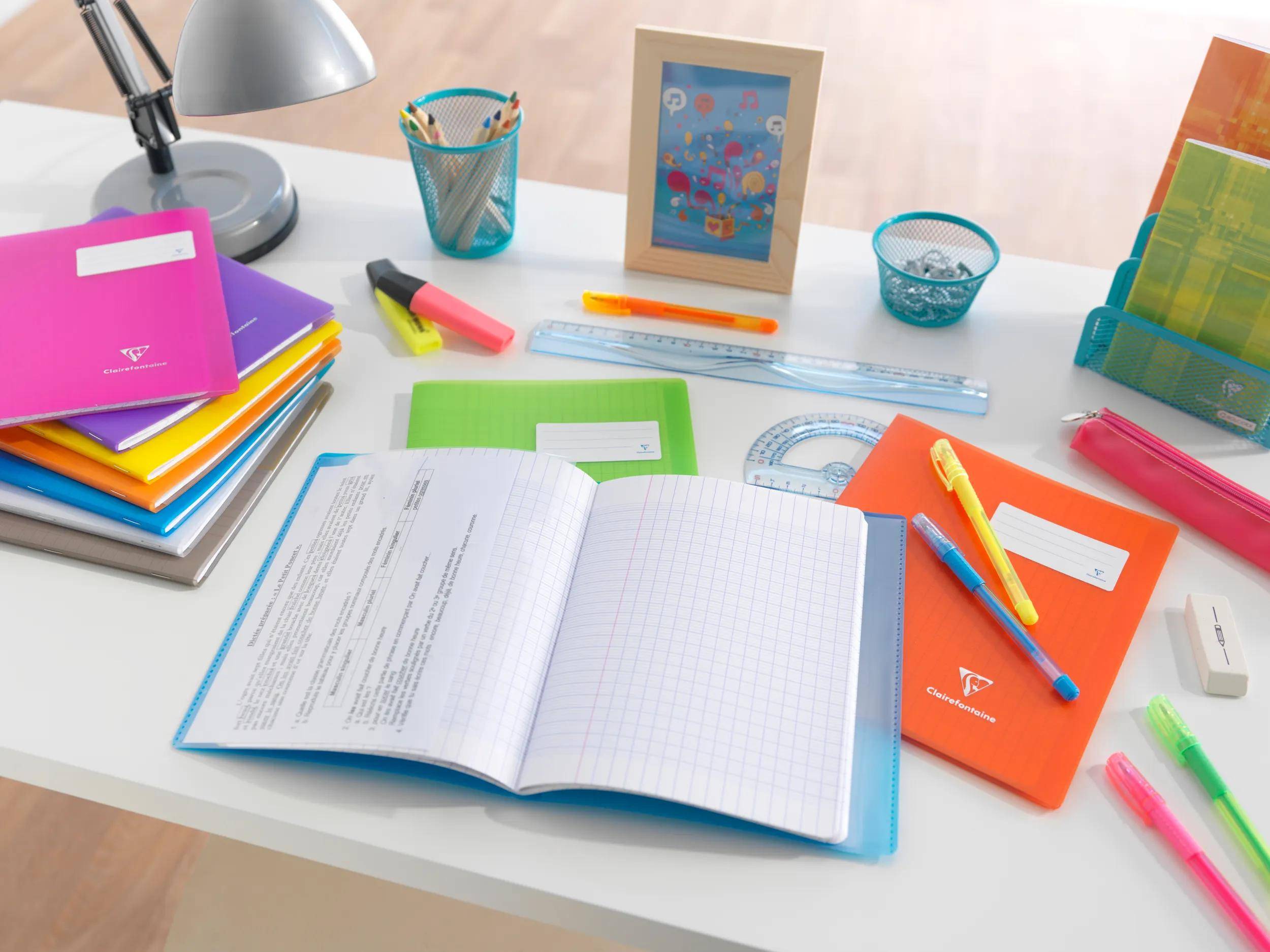 Ein ordentlicher Schreibtisch in der Schule mit einem offenen Notizbuch und verstreuter farbenfroher Schreibwaren, darunter Stifte, Textmarker, Lineale, Ordner, eine Lampe und ein Fotorahmen.