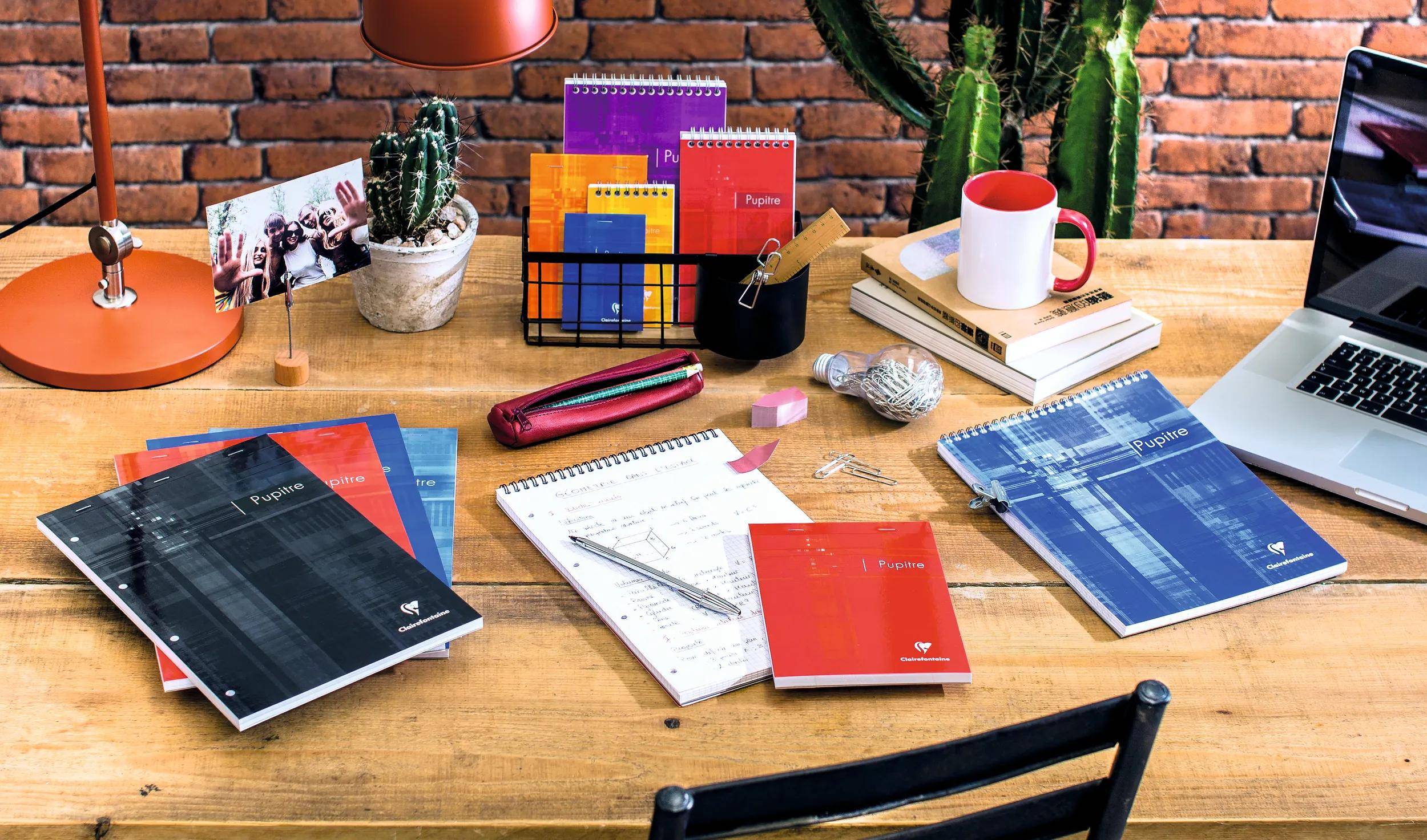 Ein hölzerner Schreibtisch mit offenen Notizbüchern, bunten Ordnern, einer roten Federmappe, Stiften, einer Tasse und Kakteen in Töpfen vor einer Ziegelwandhintergrund.