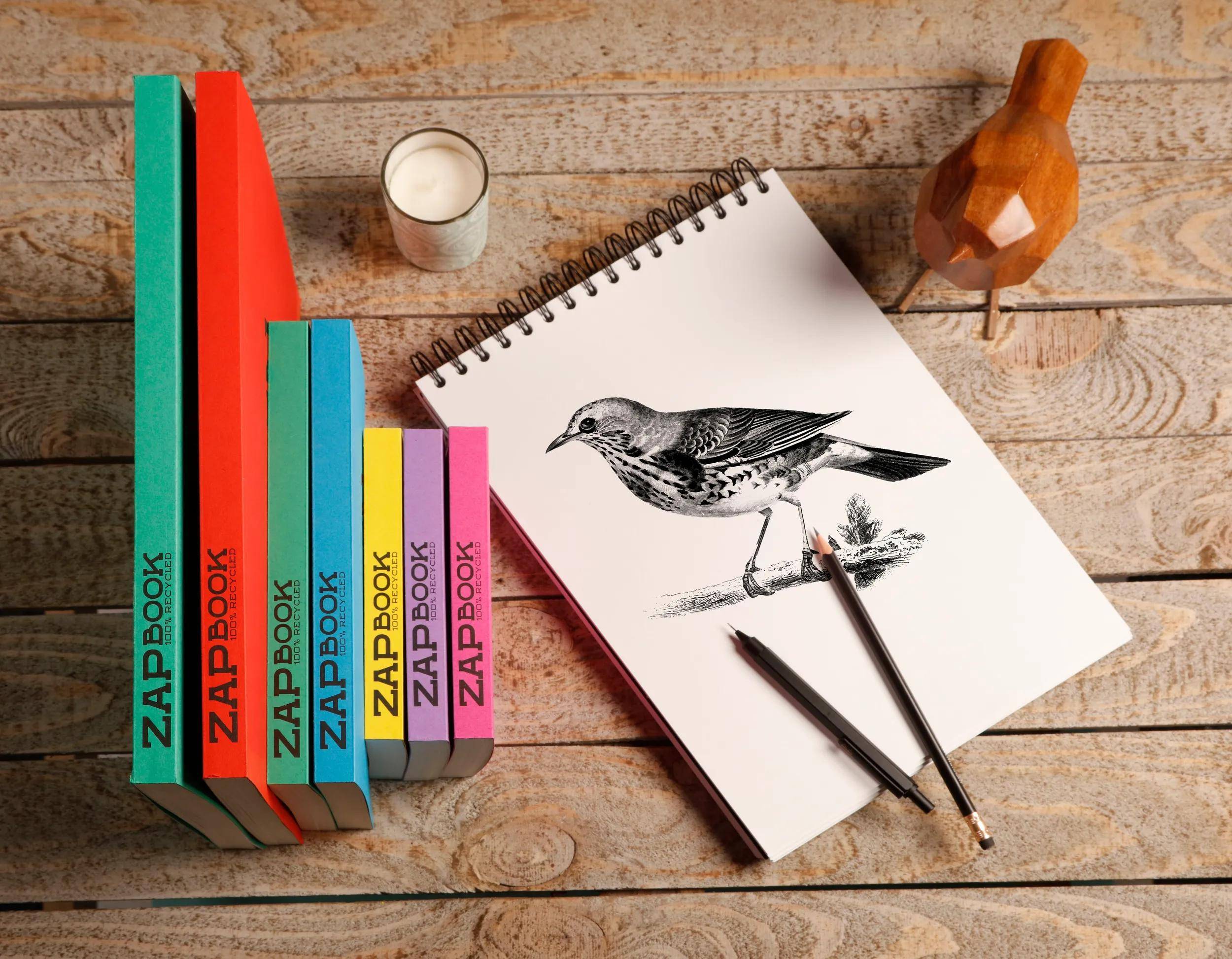 Kunstmaterialien auf einem Holztisch arrangiert neben farbenfrohen ZIPBOOK-Notizbüchern; ein Skizzenblock zeigt eine Vogelzeichnung mit Bleistiften und einer Kerze in der Nähe.