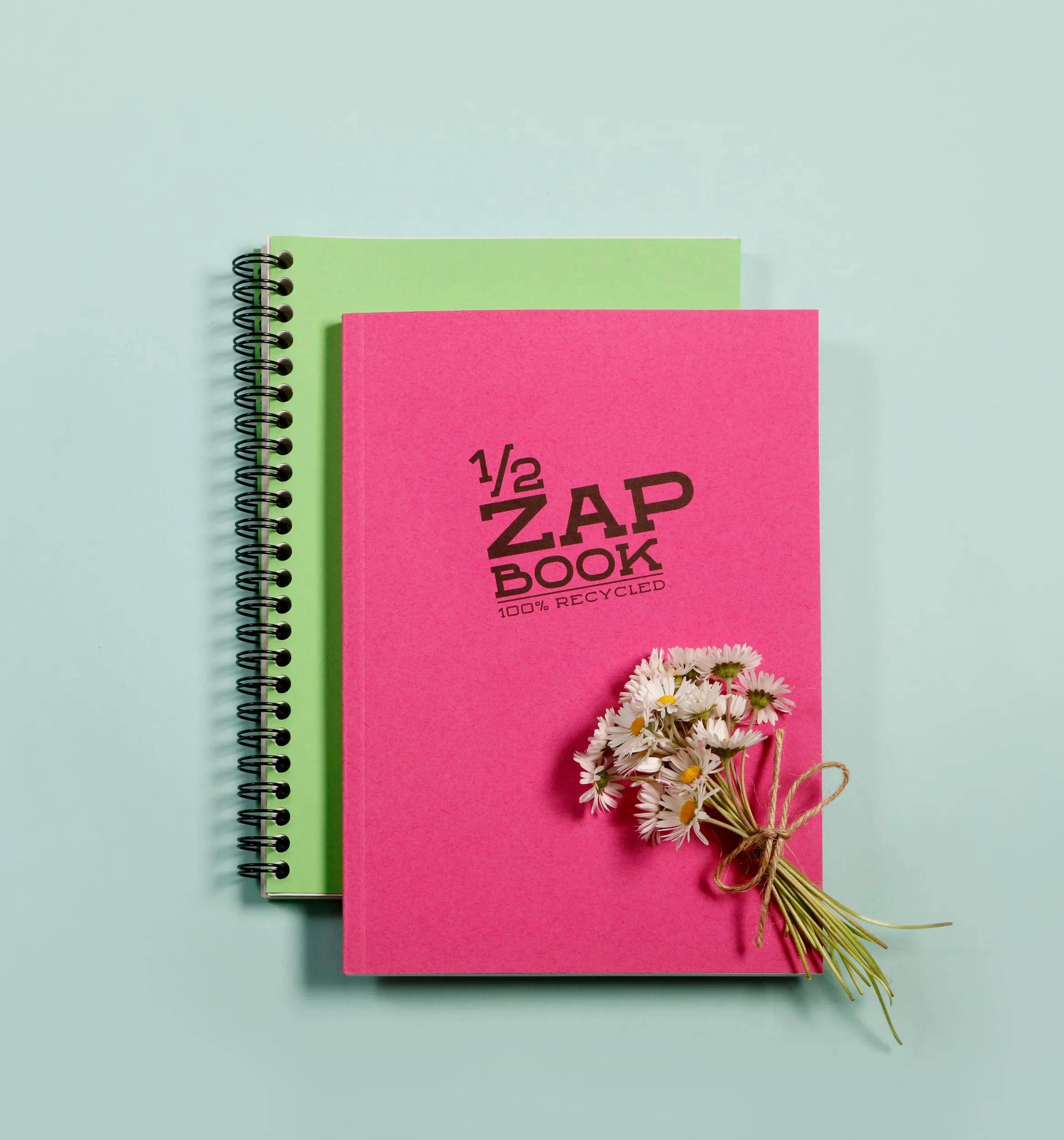 Zwei Spiralhefte, rosa vorne und grün hinten, mit einem kleinen gebundenen Gänseblümchenstrauß auf dem rosa Umschlag mit der Aufschrift „1/2 ZAP BOOK 100% RECYCLED