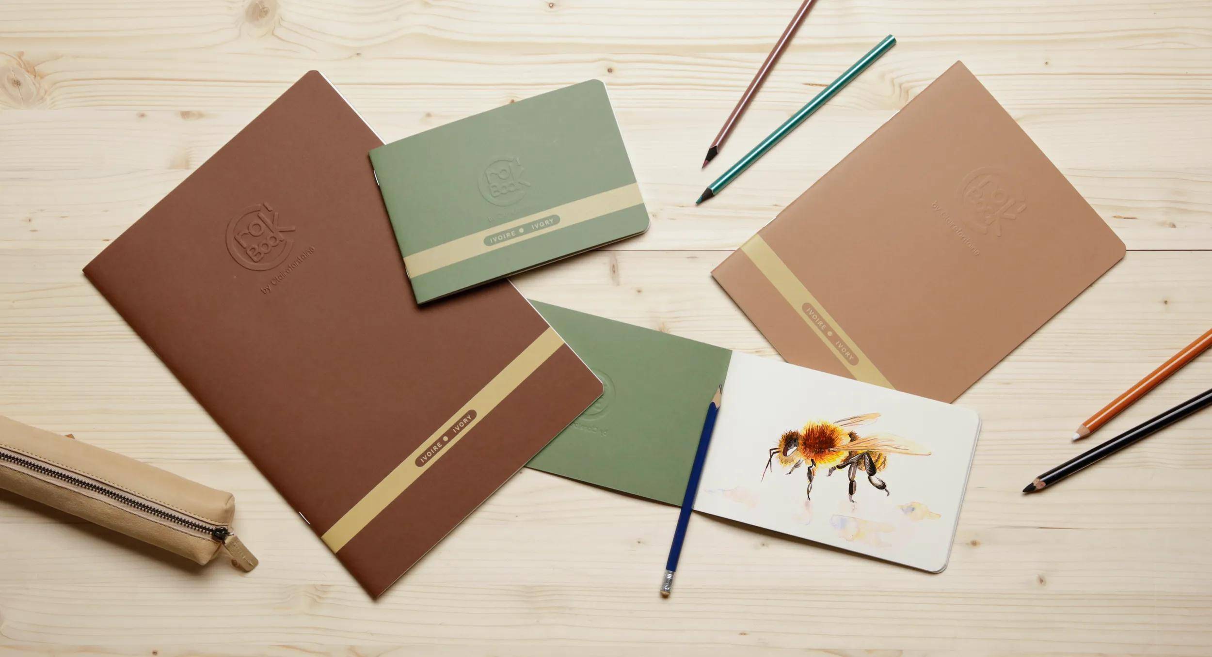 Mehrere Notizbücher und Bleistifte auf einem hellen Holzschreibtisch angeordnet, mit einer kleinen Karte, auf der eine Bienenillustration abgebildet ist.