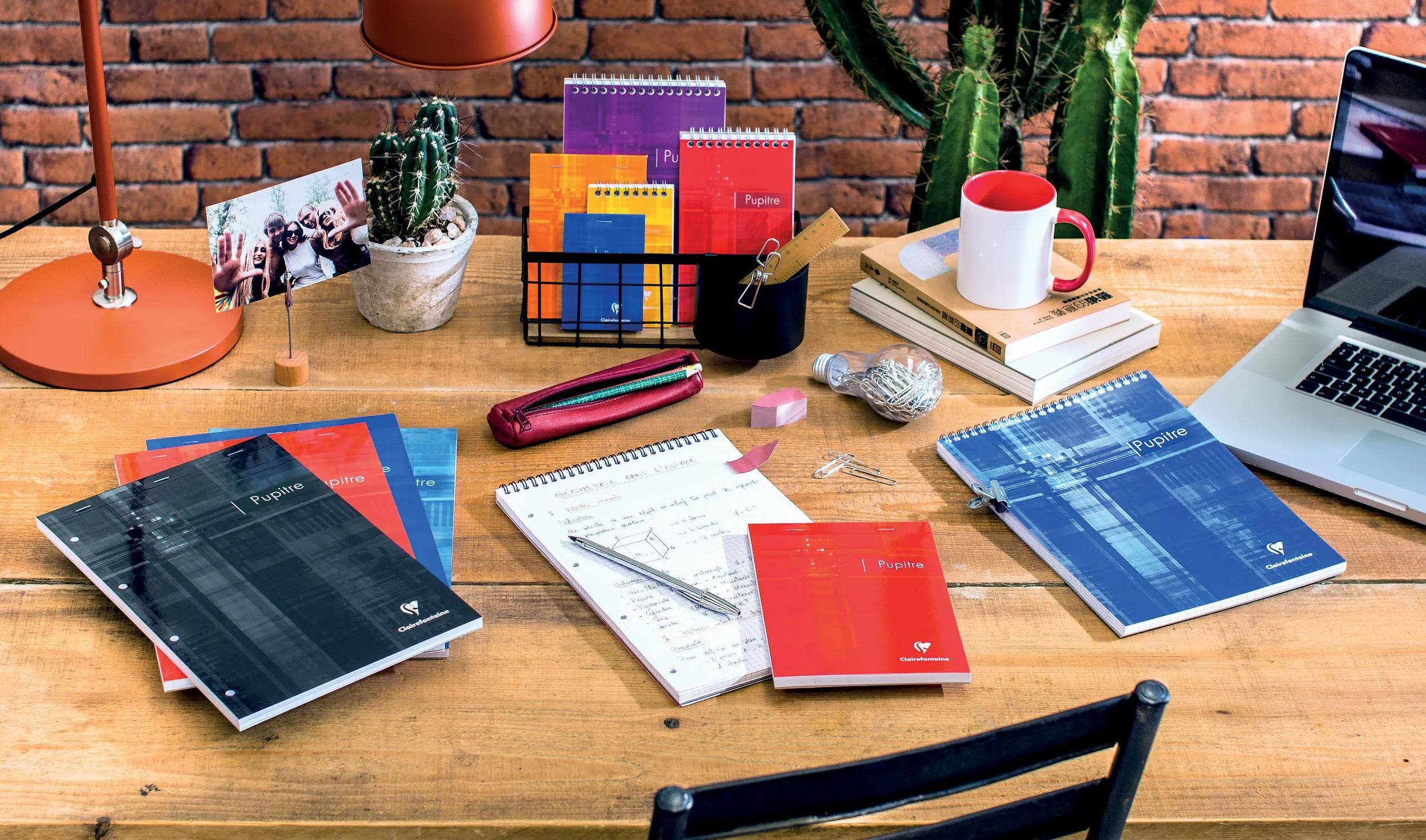 Schreibtisch mit gestapelten Notizbüchern, einem Planer, Stiften, Büchern, einer Kaffeetasse, einem Laptop, einem Kaktus und einer Schreibtischlampe in einem Home-Office-Setting.