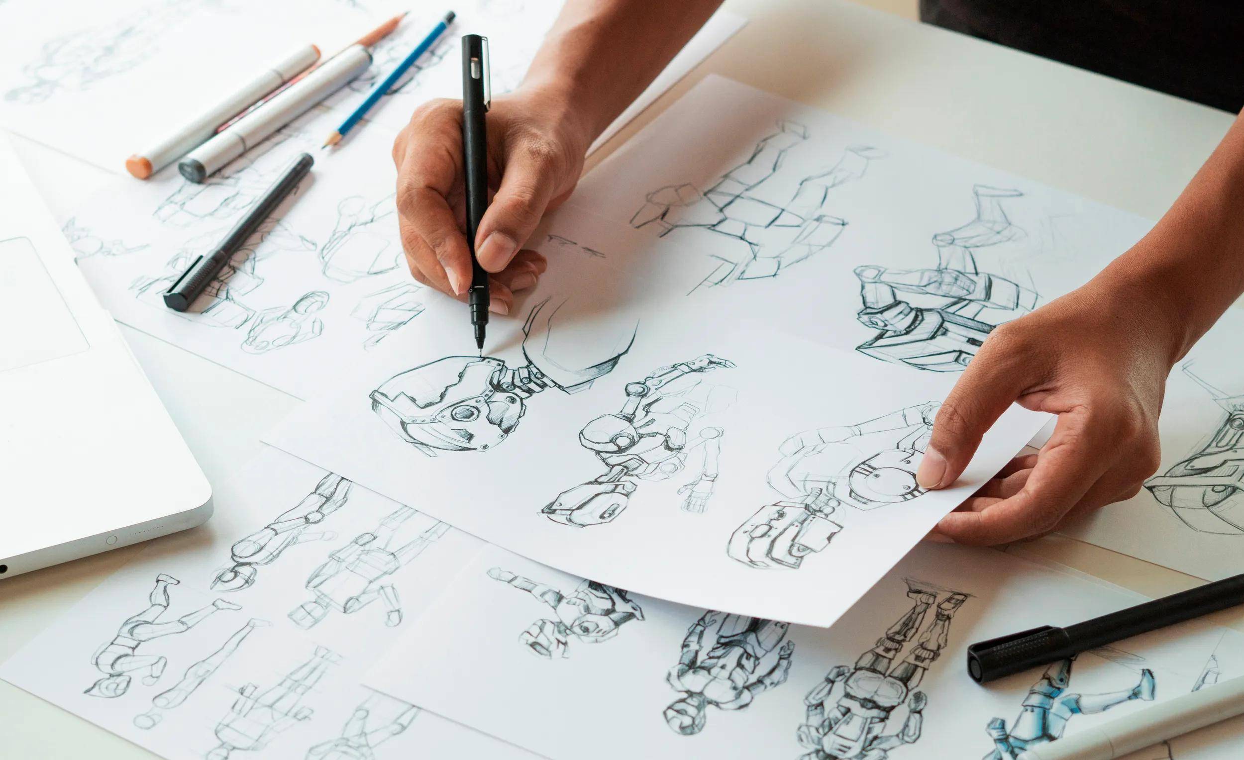 Hände, die Roboter-Charakterdesigns auf Papier mit Bleistiften und einem Laptop in der Nähe skizzieren.