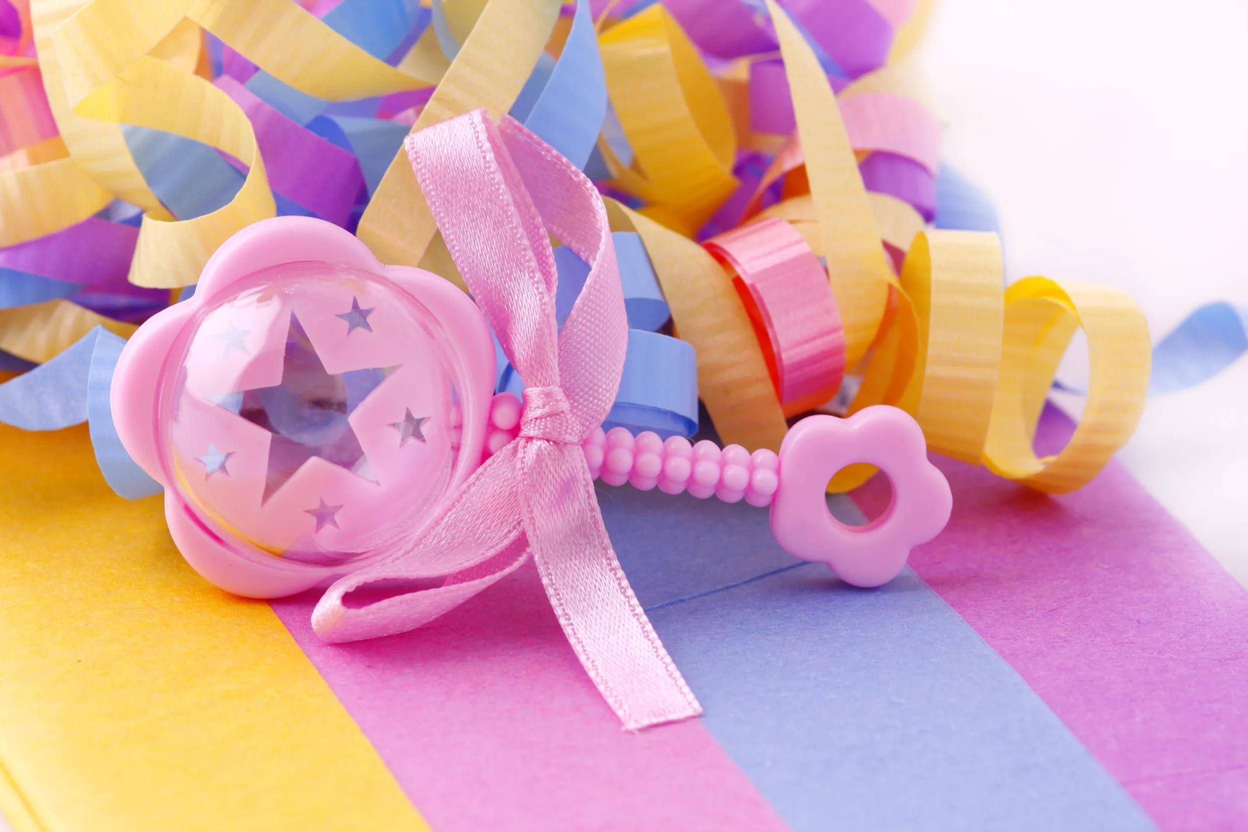 Rosa Babyrassel mit einem Band liegt auf pastellfarbigem gestreiftem Papier inmitten von bunten gekräuselten Partyschleifen.