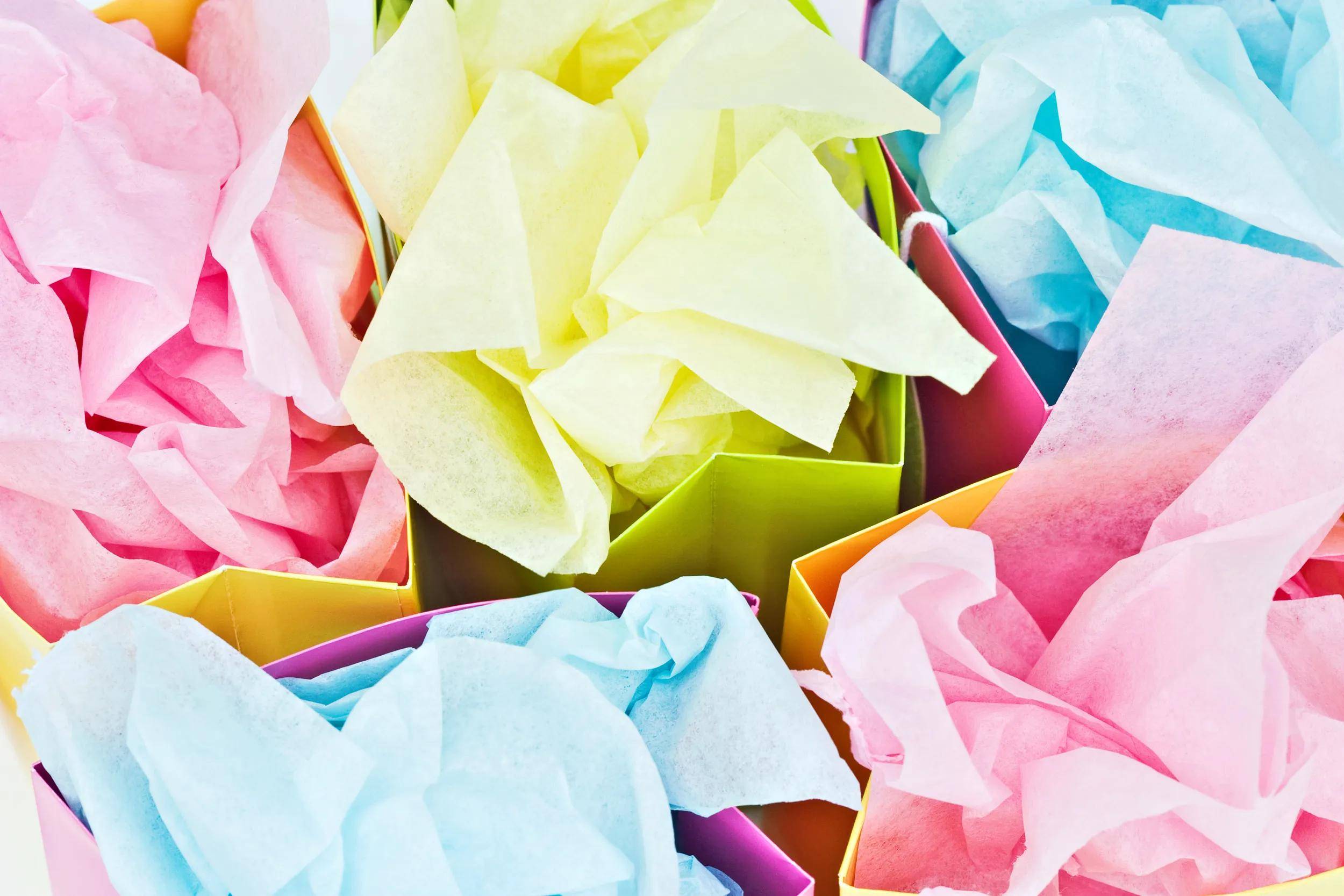 Bunte Pastellseidenpapiere, die in mehrere offene Geschenkboxen gefaltet sind und den Rahmen in Rosa, Gelb, Blau und Orange ausfüllen.